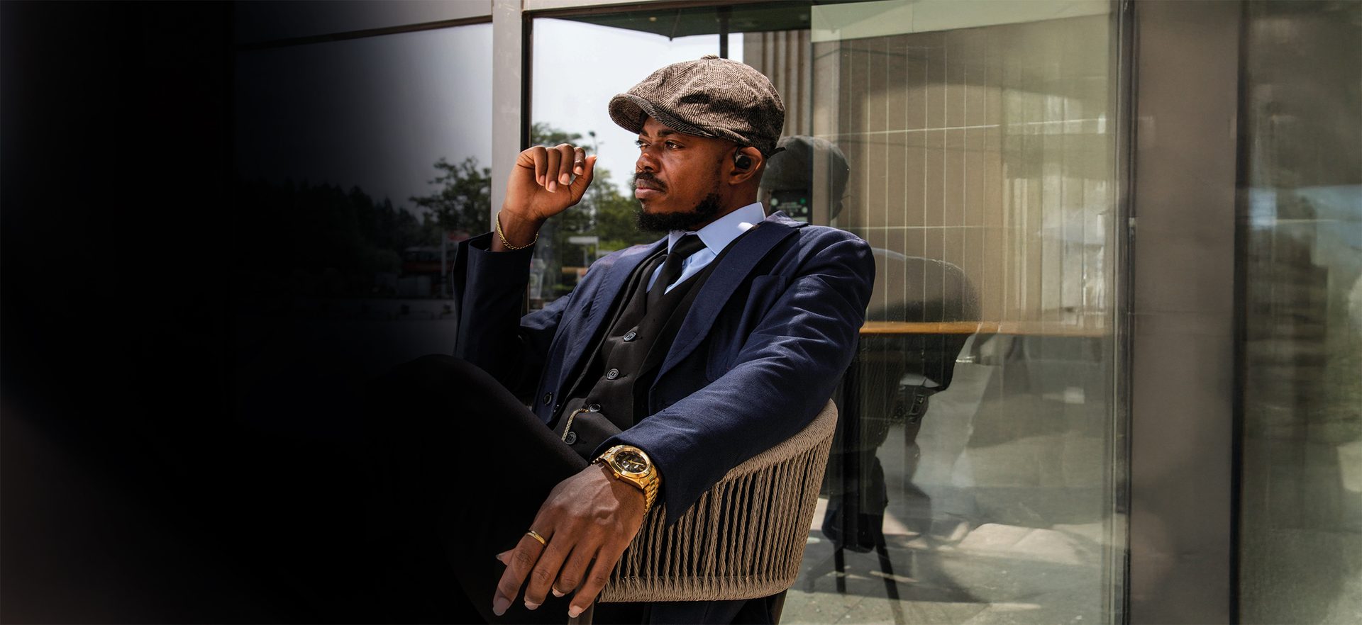 Stylish man in a suit, flat cap, and earbuds, wearing a gold watch, sitting outdoors.