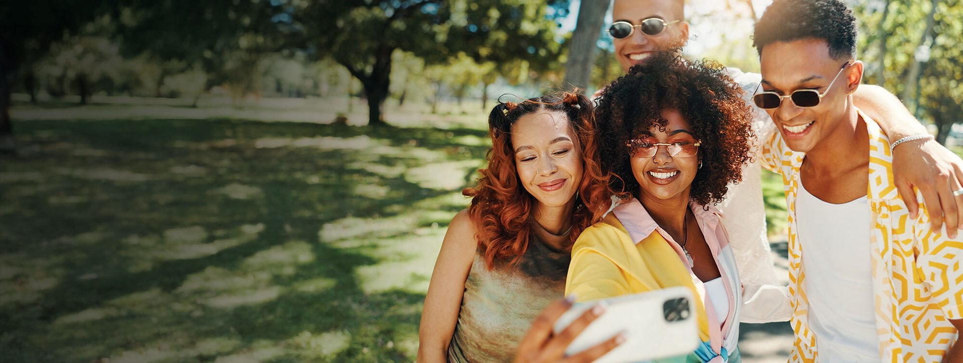 Four diverse friends smiling and taking a selfie in a sunny park.