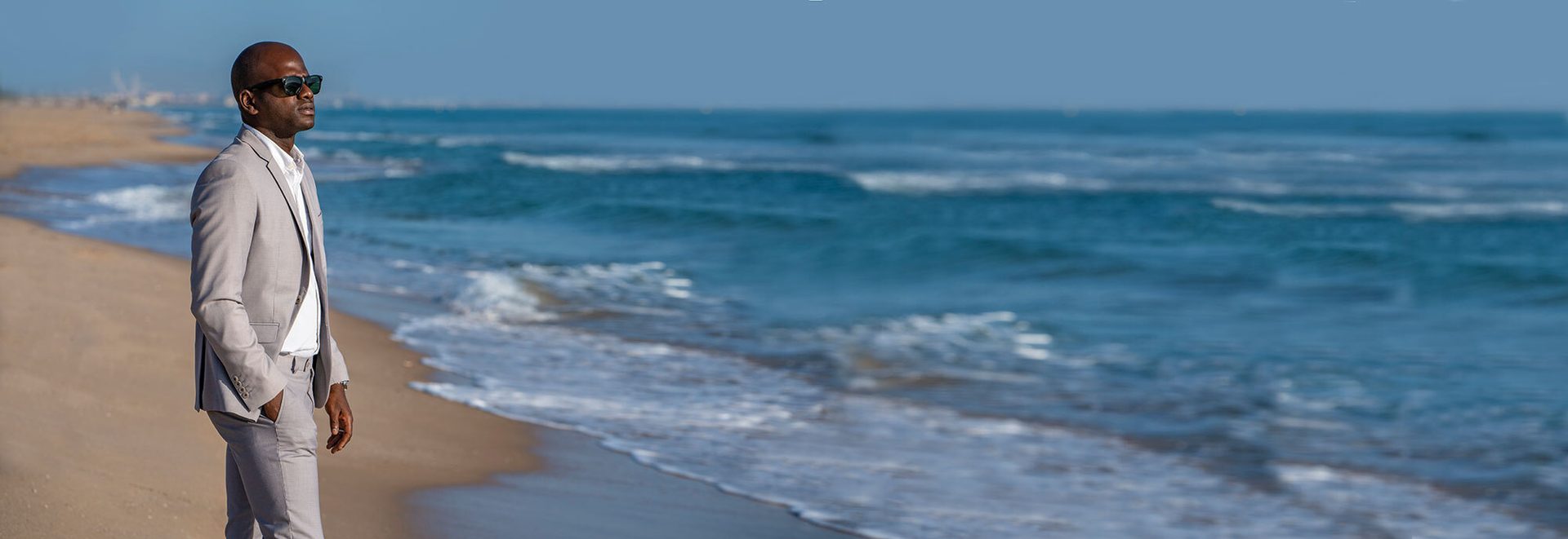 A man in a light suit and sunglasses stands on a sandy beach next to the ocean.