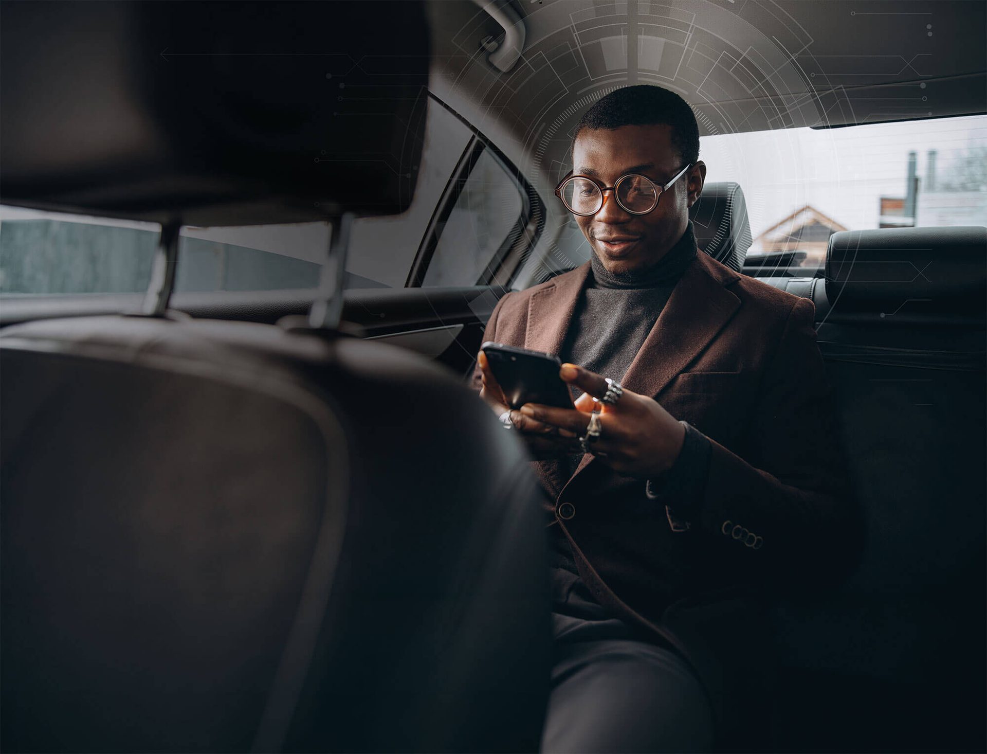 A stylish man in glasses and a brown coat smiles while looking at his smartphone in the back of a car.
