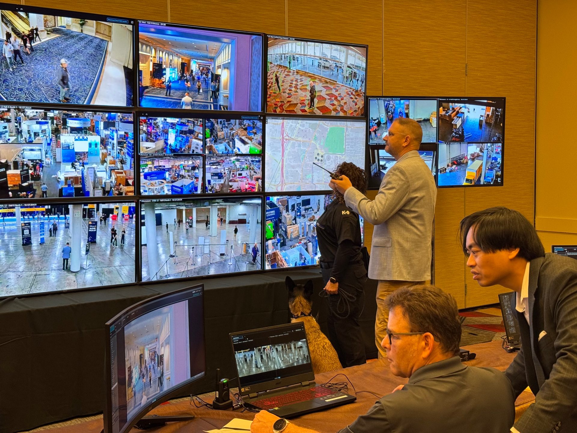 Security personnel and a K-9 dog monitor a large video wall displaying multiple surveillance feeds.