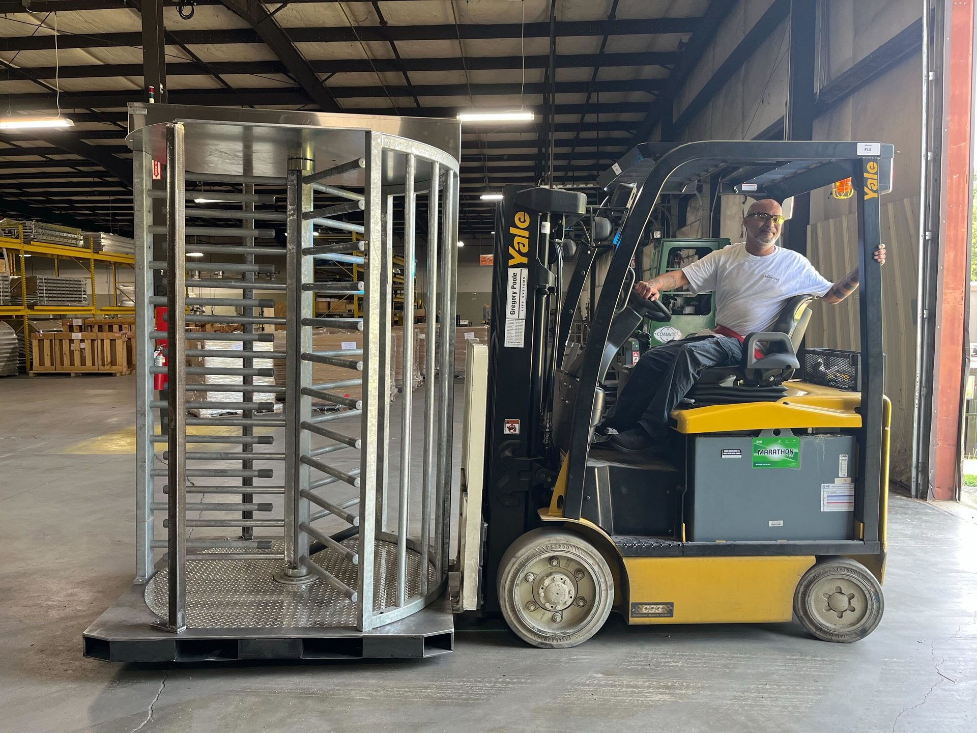 A man on a yellow forklift next to a large stainless steel turnstile in a warehouse.