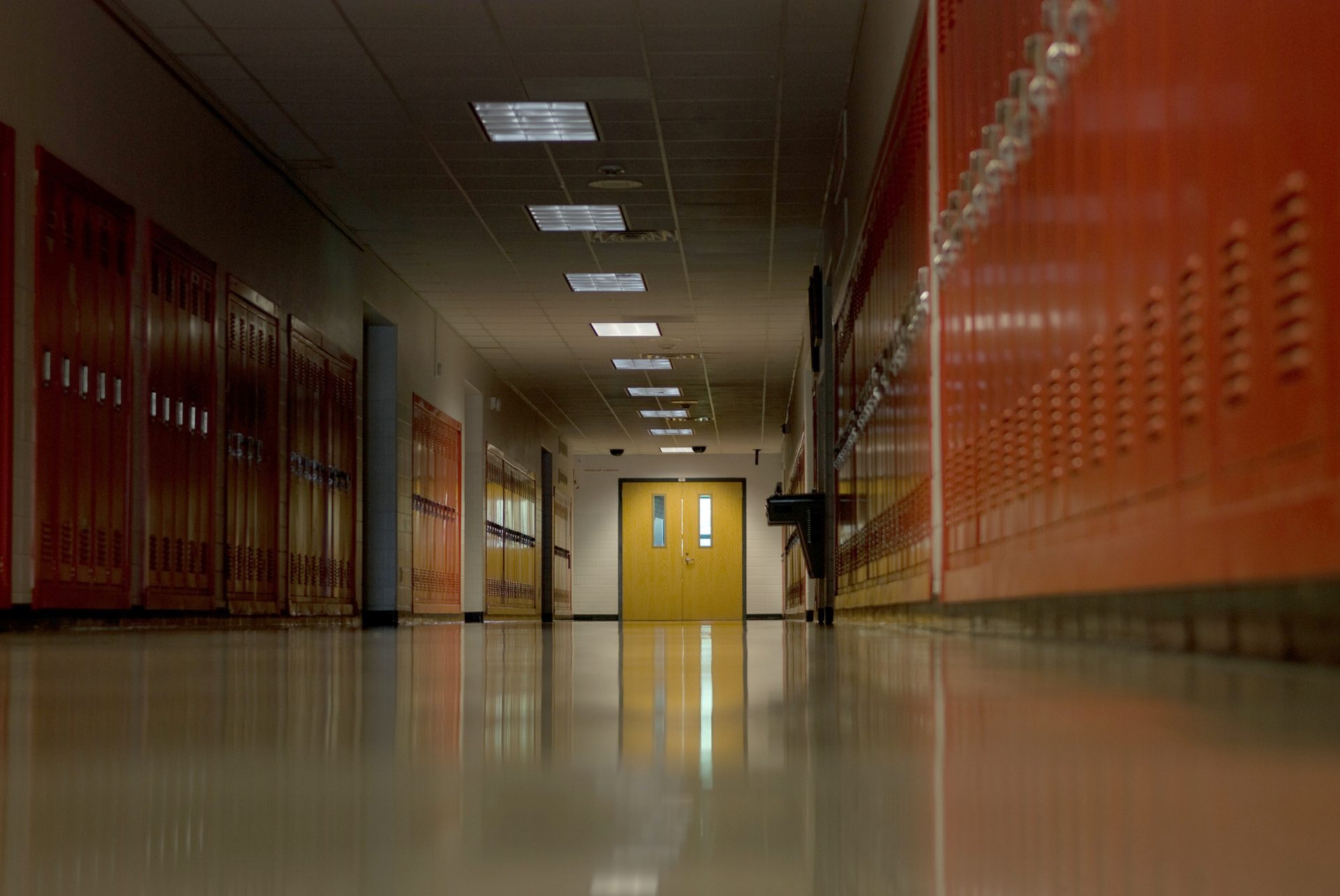 Long, empty school hallway with orange lockers and a reflective floor.