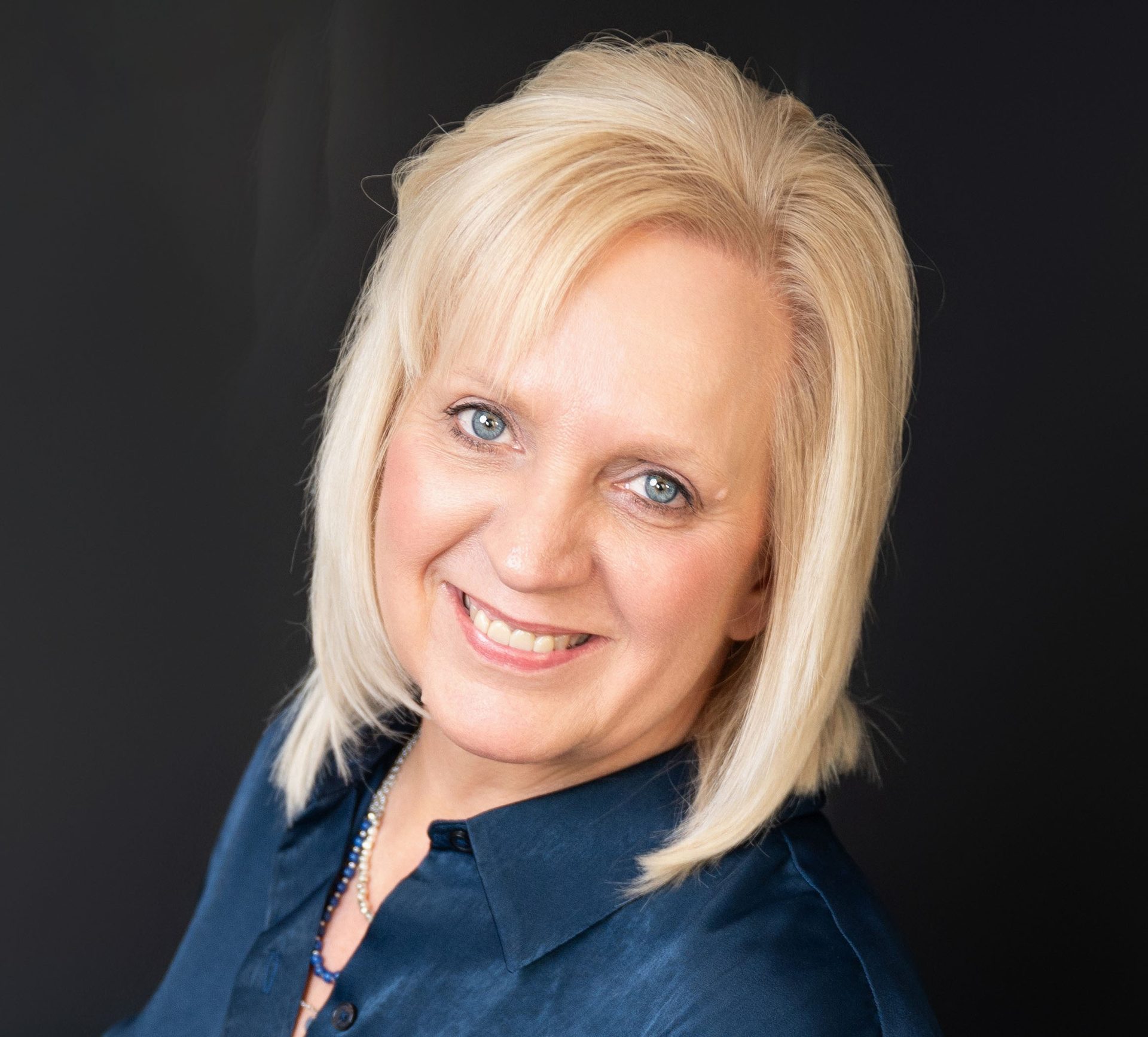 Smiling blonde woman with blue eyes in a blue shirt against a dark background.