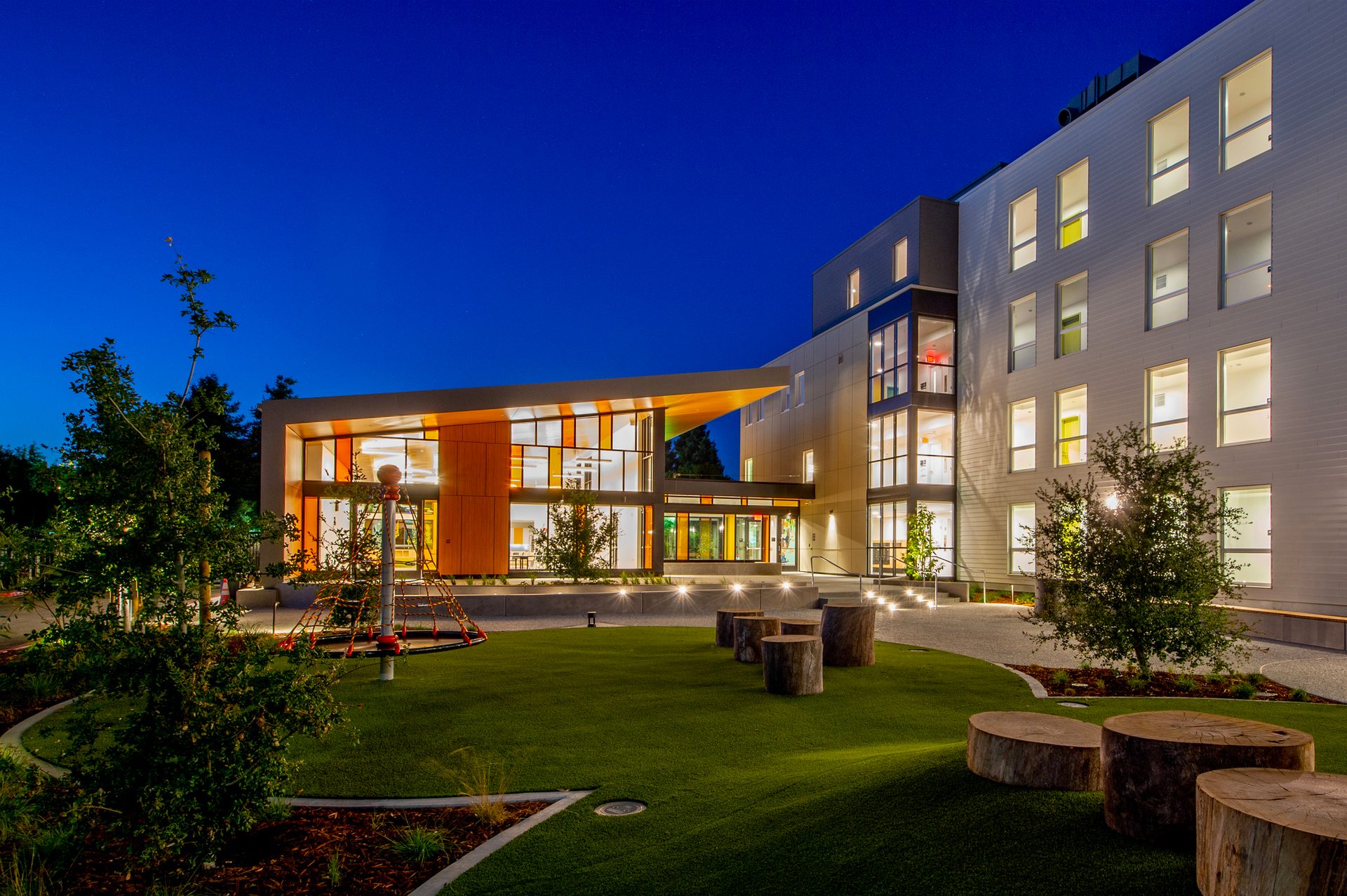 Modern building at dusk, illuminated interiors, green lawn, tree stumps, and play structure.