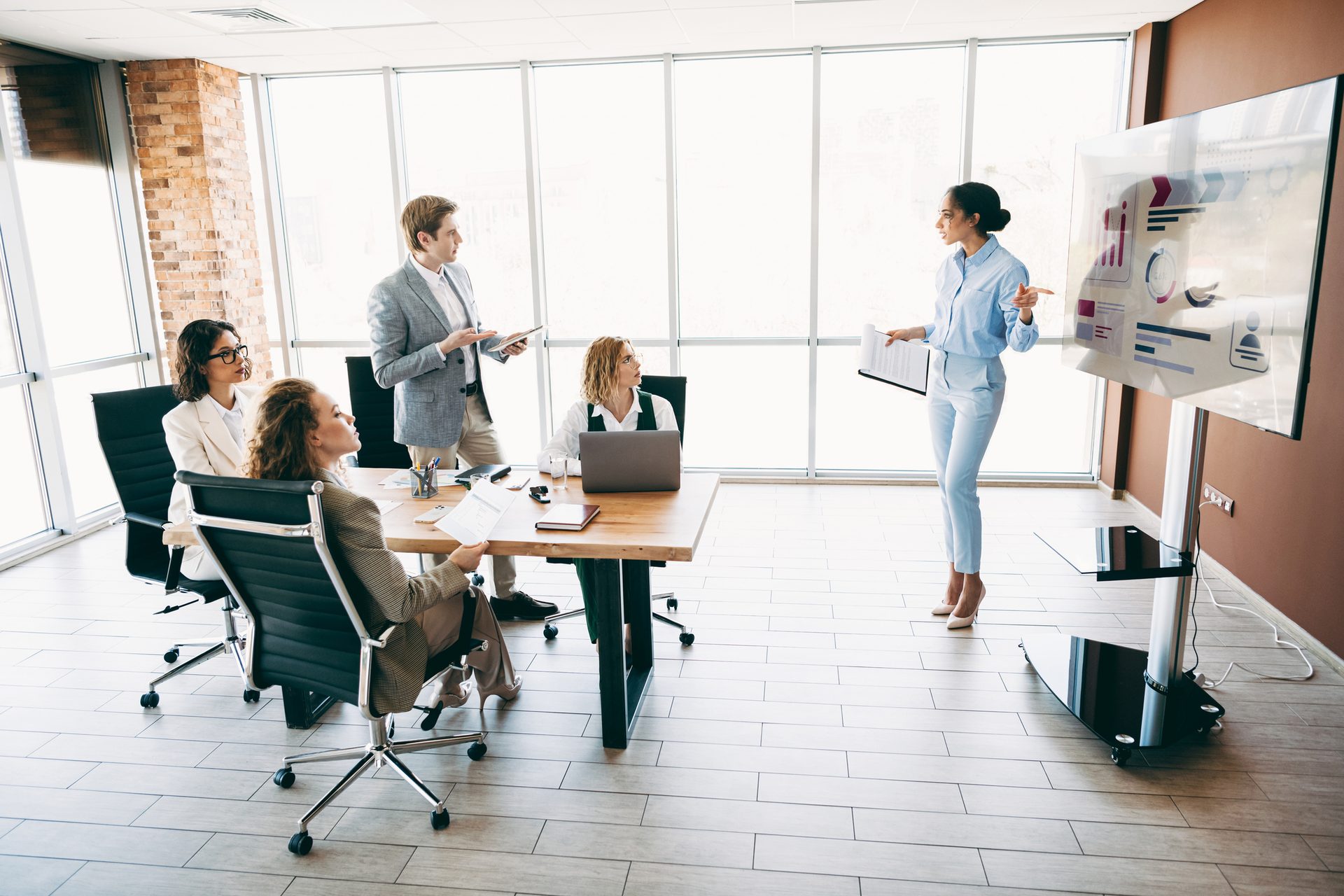 Five business professionals in a bright modern office meeting. A woman presents data on a large screen.
