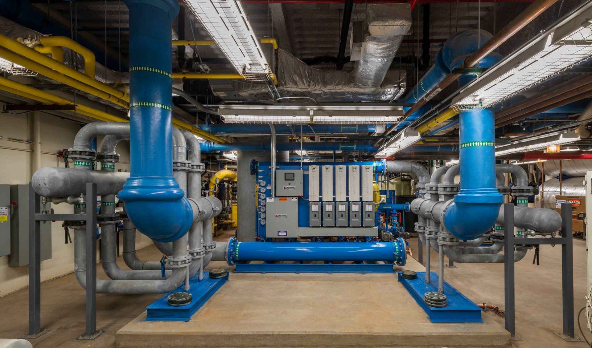 Mechanical room with intricate blue, grey, and yellow industrial pipes and large central machinery.