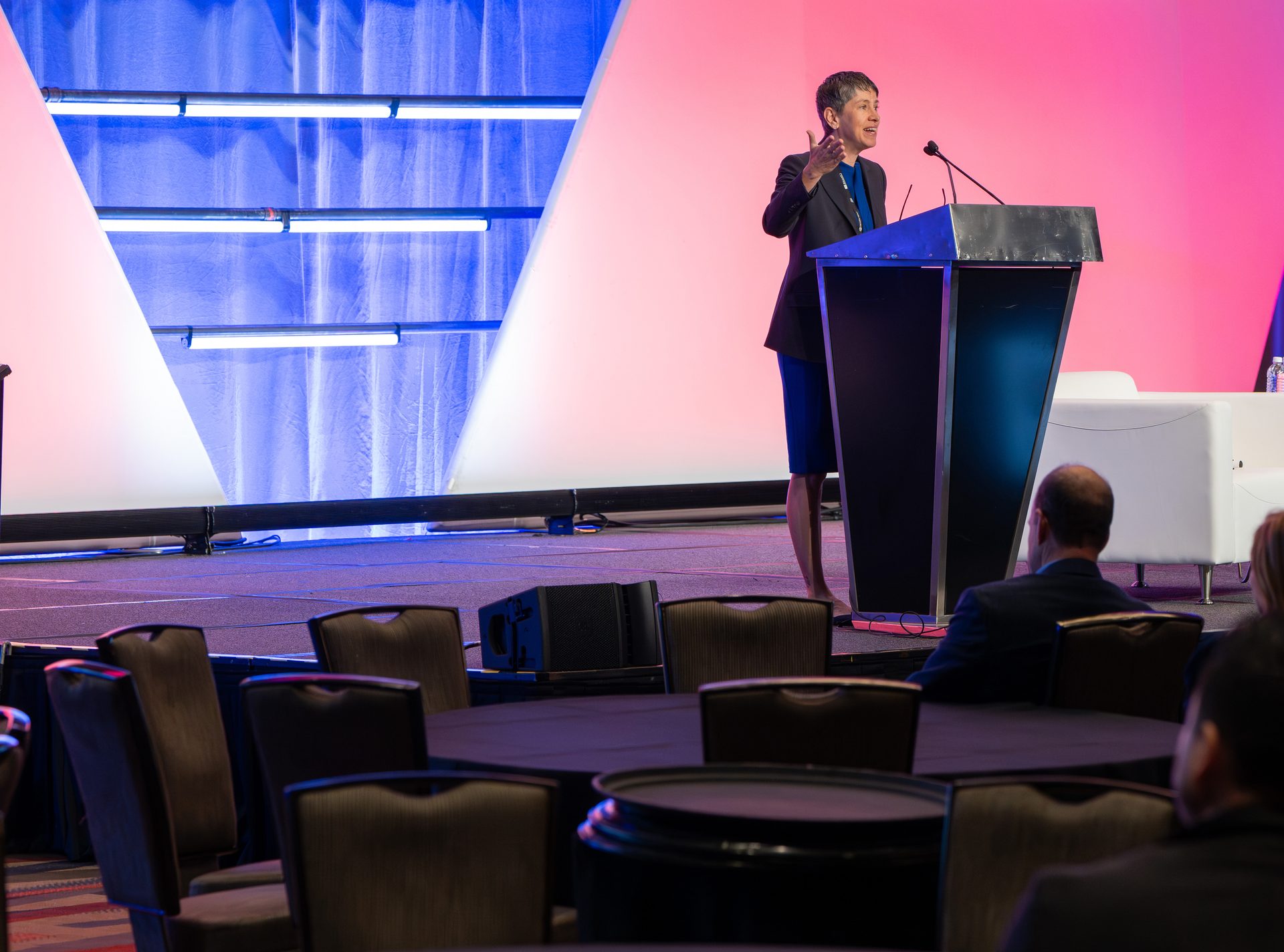 Woman addresses an audience from a podium on a stage with vibrant lighting.
