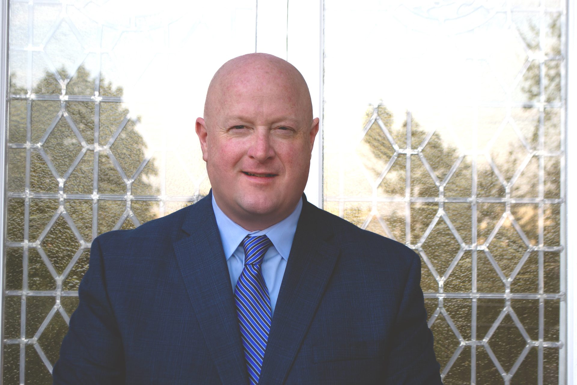 Bald man in a blue suit and striped tie smiles in front of a patterned glass door.