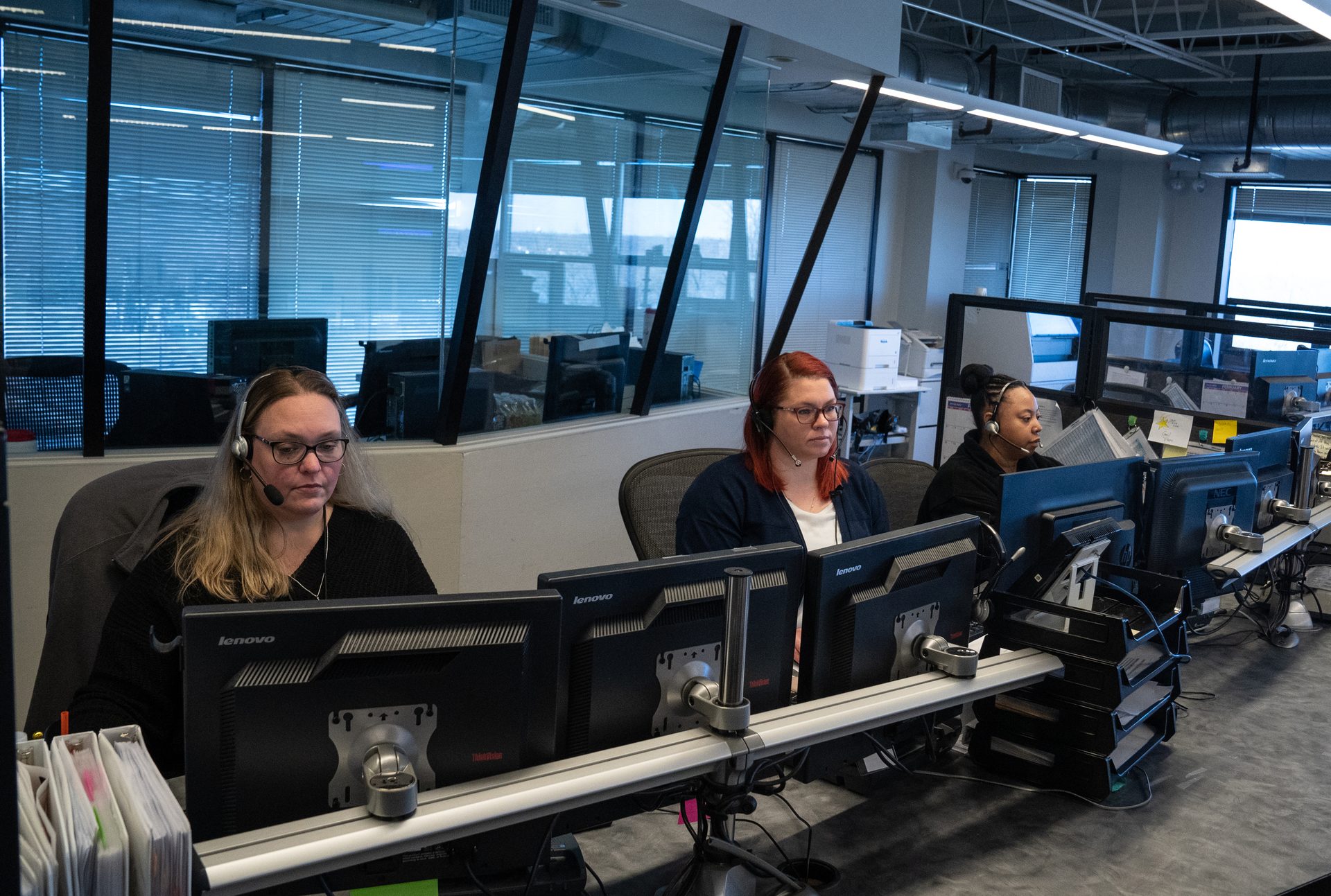 Three women with headsets work at computer stations in an office.