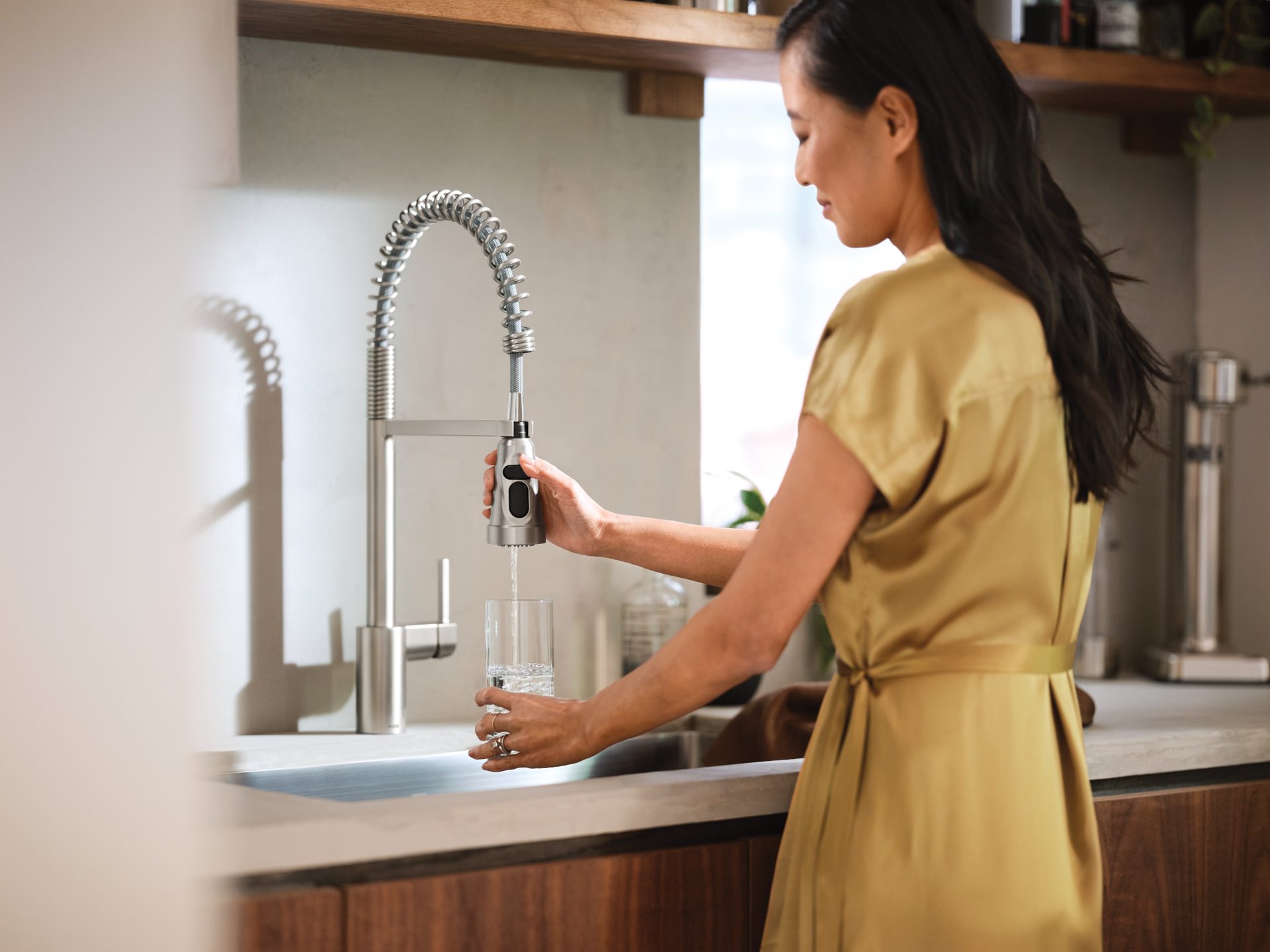 A woman in a gold dress fills a glass of water from a modern kitchen faucet.