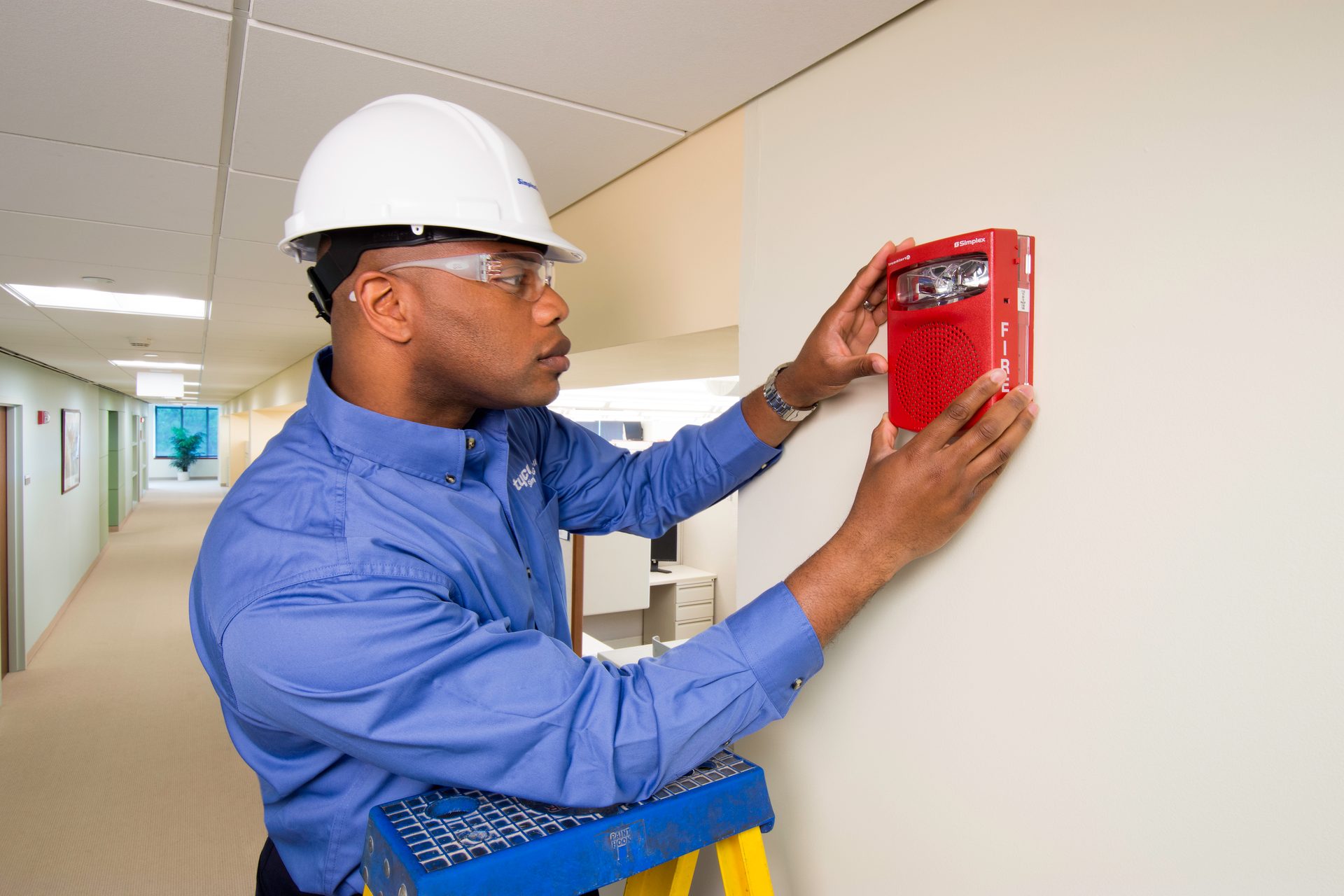 A man in safety gear installs a red fire alarm on a wall, standing on a step stool in a hallway.