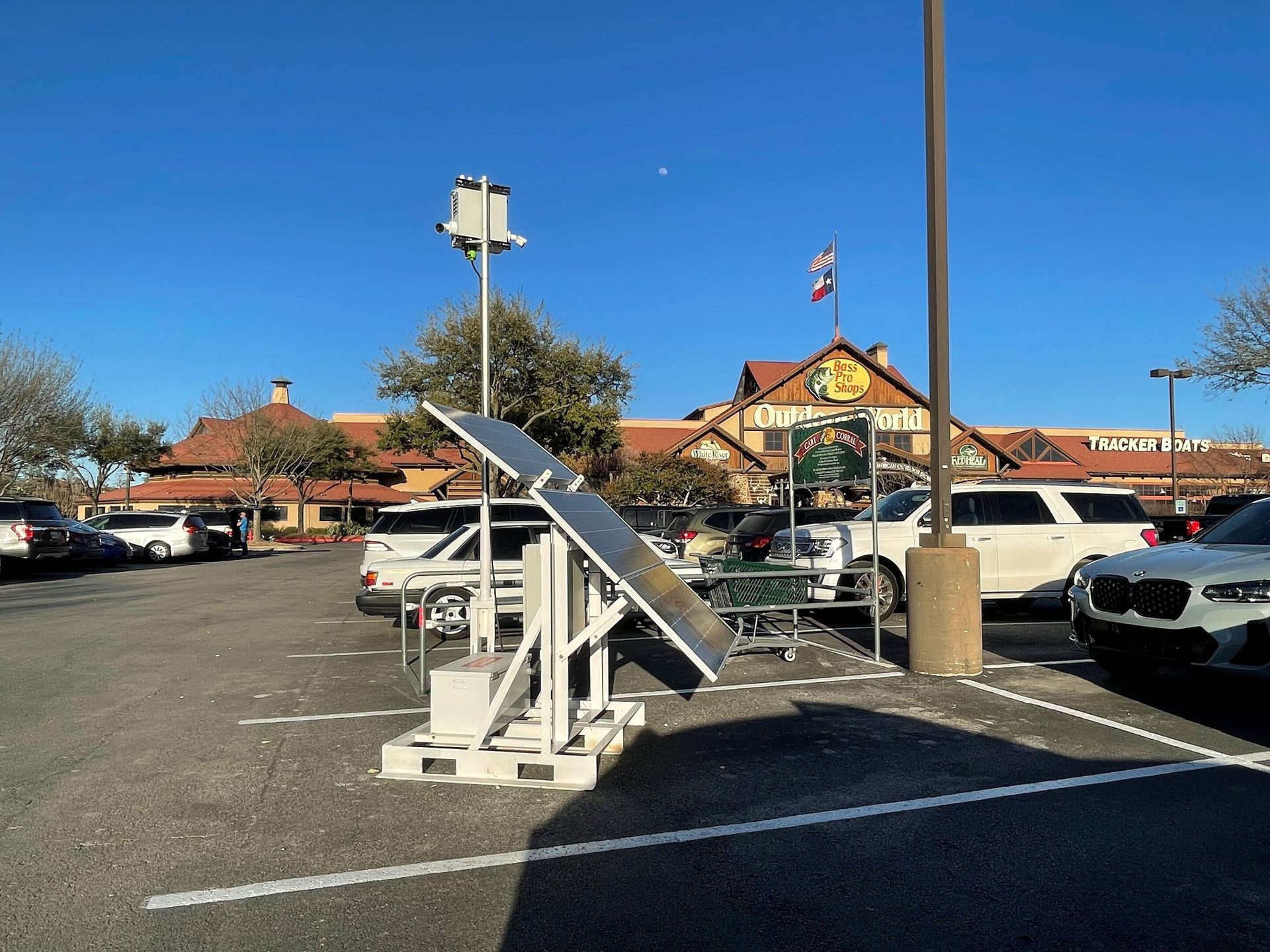 Solar-powered camera tower in a parking lot with Bass Pro Shops Outdoor World in the background under a blue sky.
