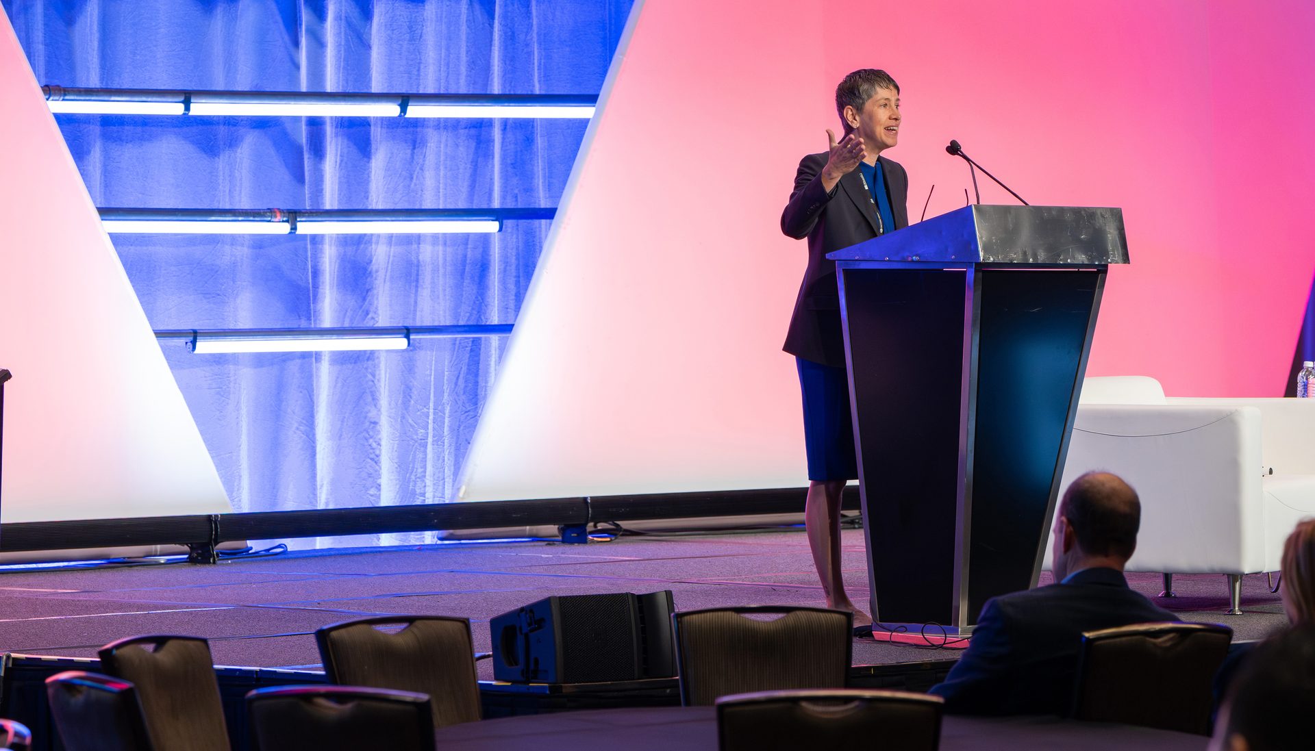 A woman in a dark suit speaks at a podium on a stage with vibrant blue and pink backlighting.