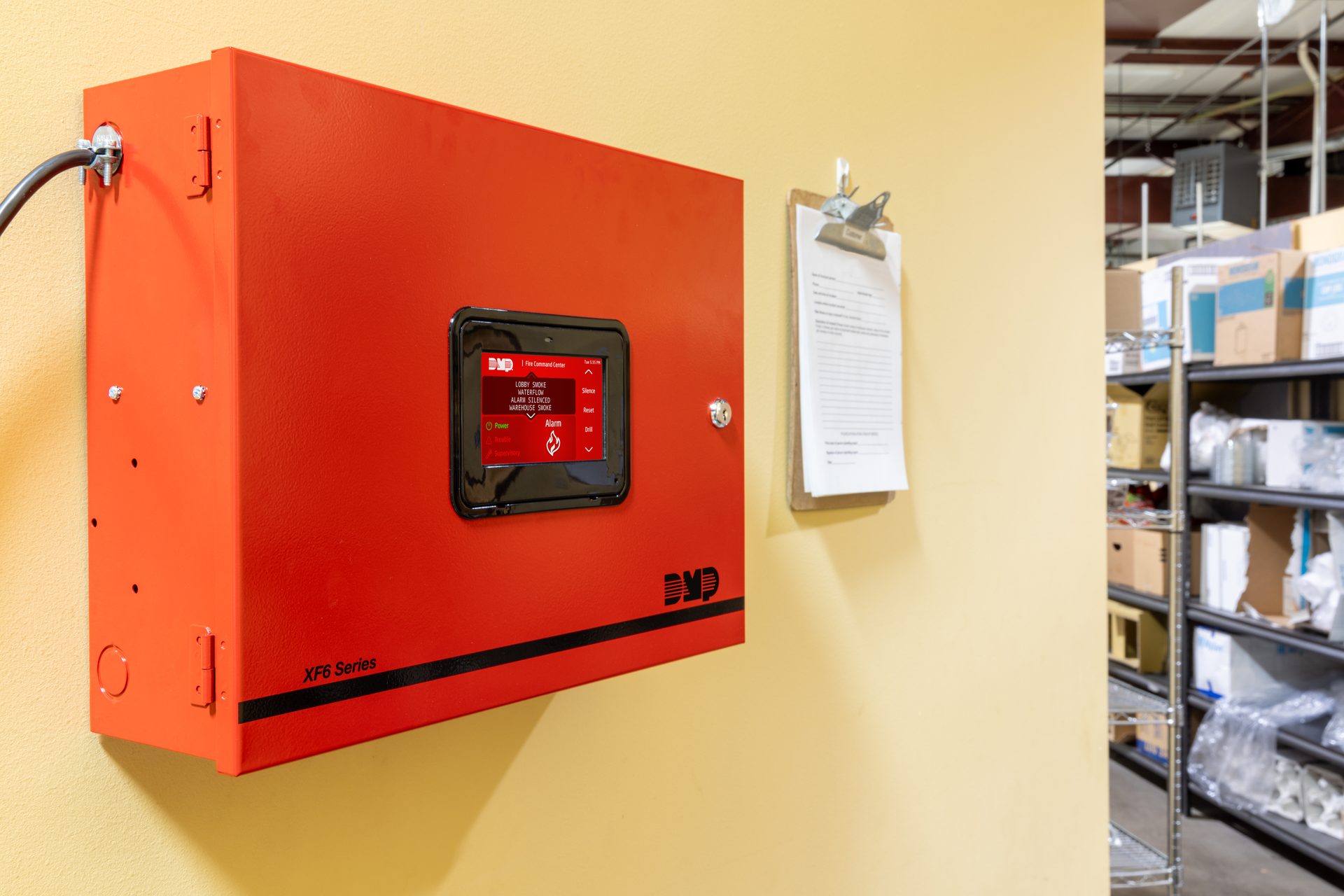 A red DMP XF6 Series fire alarm control panel with a digital display showing an active alarm, in a warehouse.