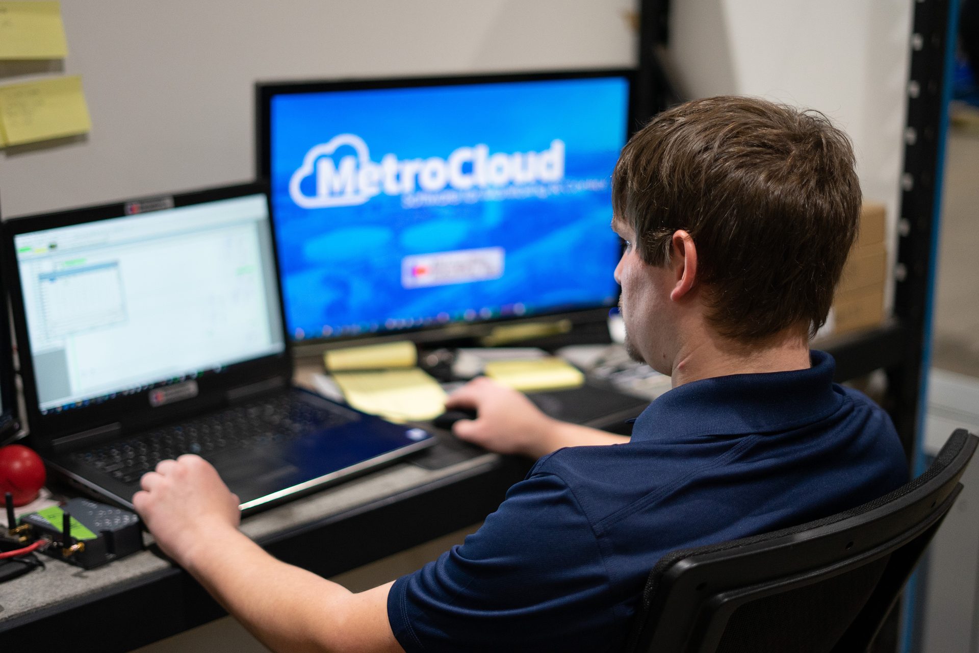 Man at a desk using a laptop and mouse, with "MetroCloud" on the main monitor.
