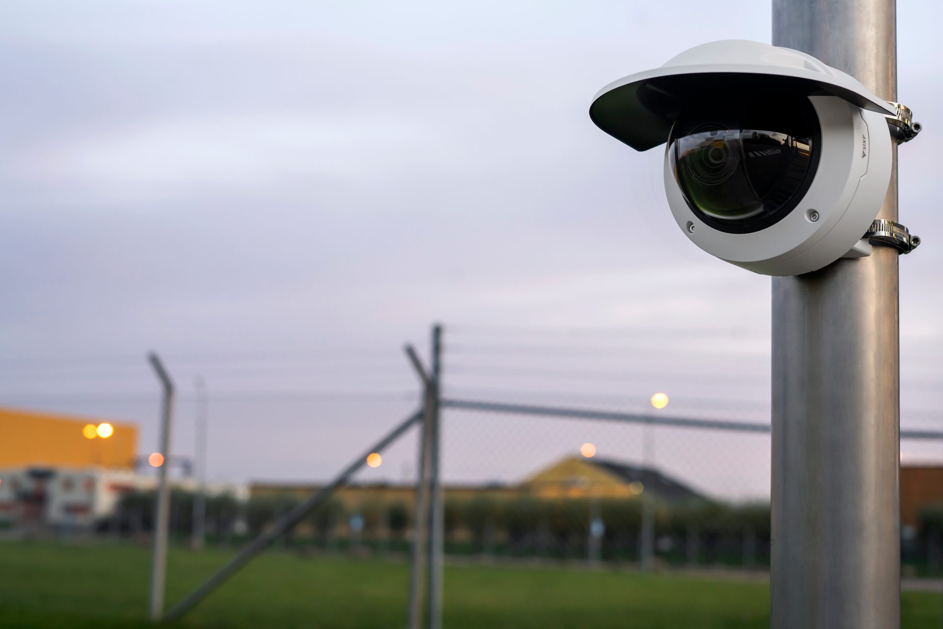 White security camera on pole, blurry outdoor fenced area in background.