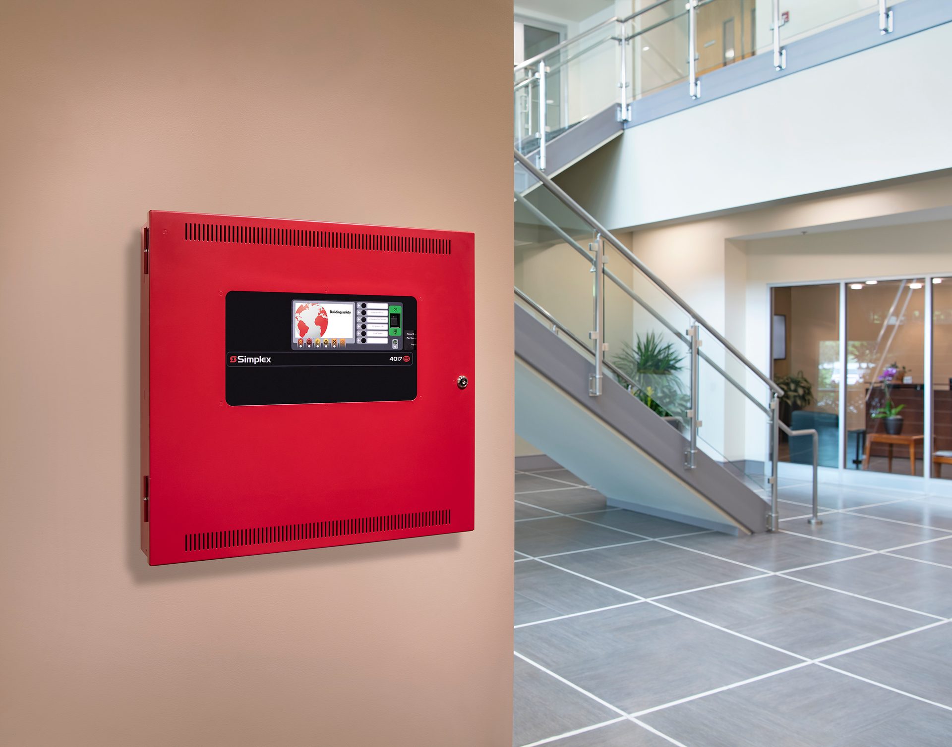 Red fire alarm panel on a wall, beside a modern building lobby with a staircase and tiled floor.
