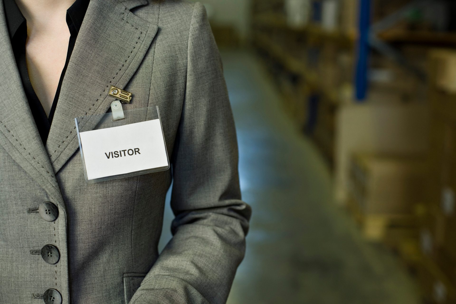 A person in a grey blazer wears a "VISITOR" badge, with a blurred warehouse in the background.