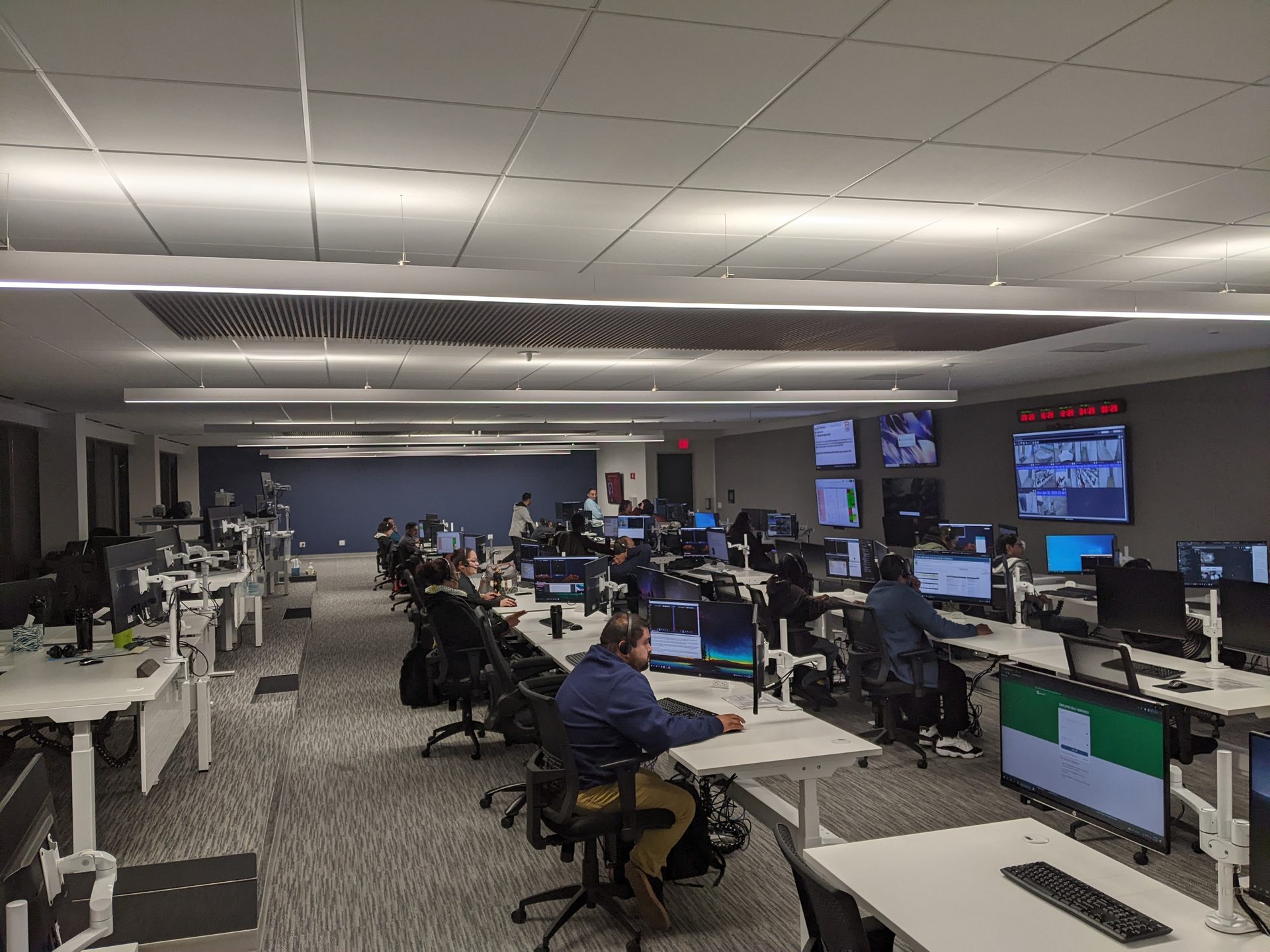People working in a modern control room or open-plan office with multiple monitors and large wall displays.