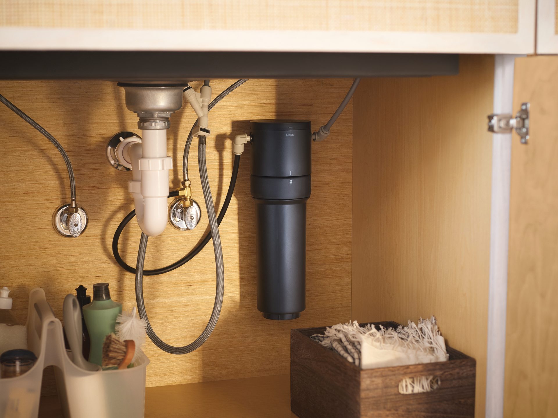 Undersink cabinet with Moen water filter, plumbing, cleaning supplies, and cloths.