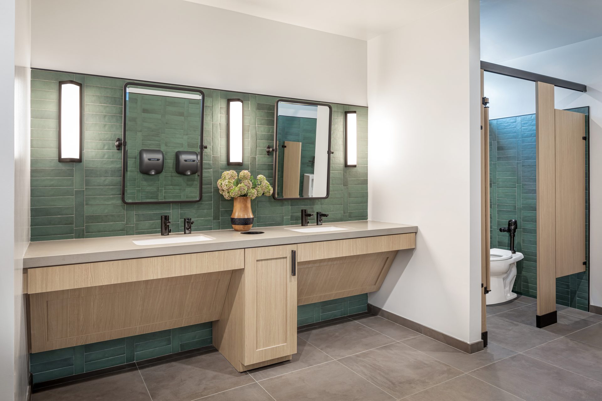 Modern restroom: green tiled walls, wood vanity, sinks, mirrors, lights, toilet stalls.