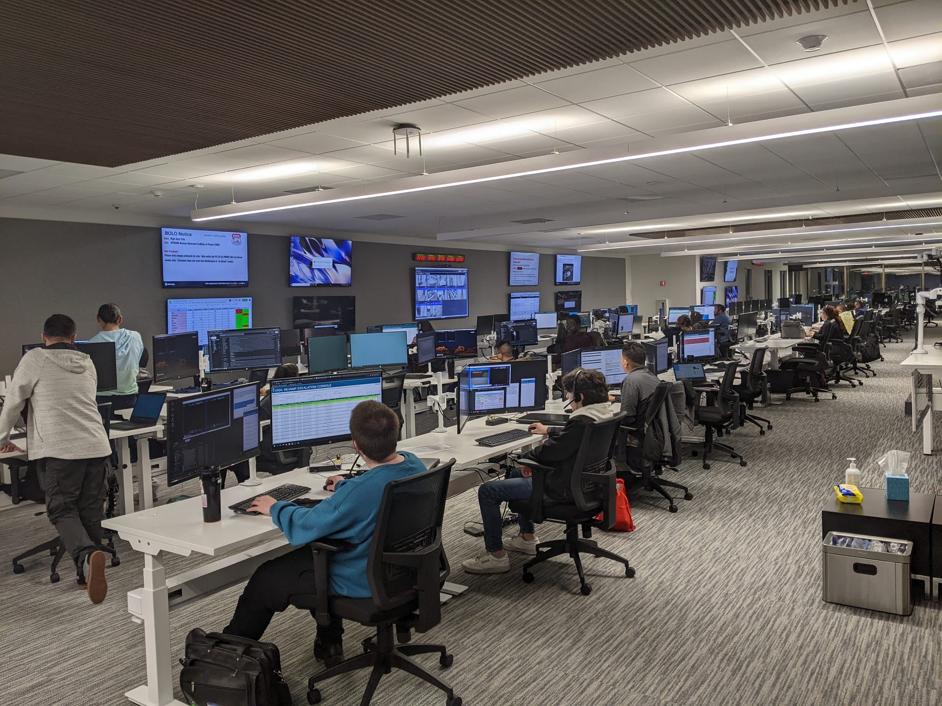 Busy open-plan office with many people working at computer workstations and large wall displays.
