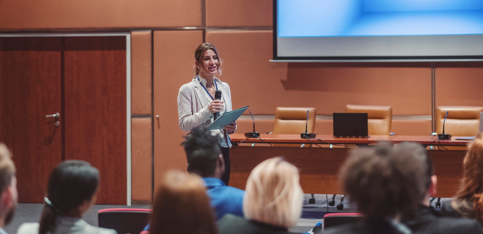 A smiling woman speaks into a microphone at a conference, addressing an audience.