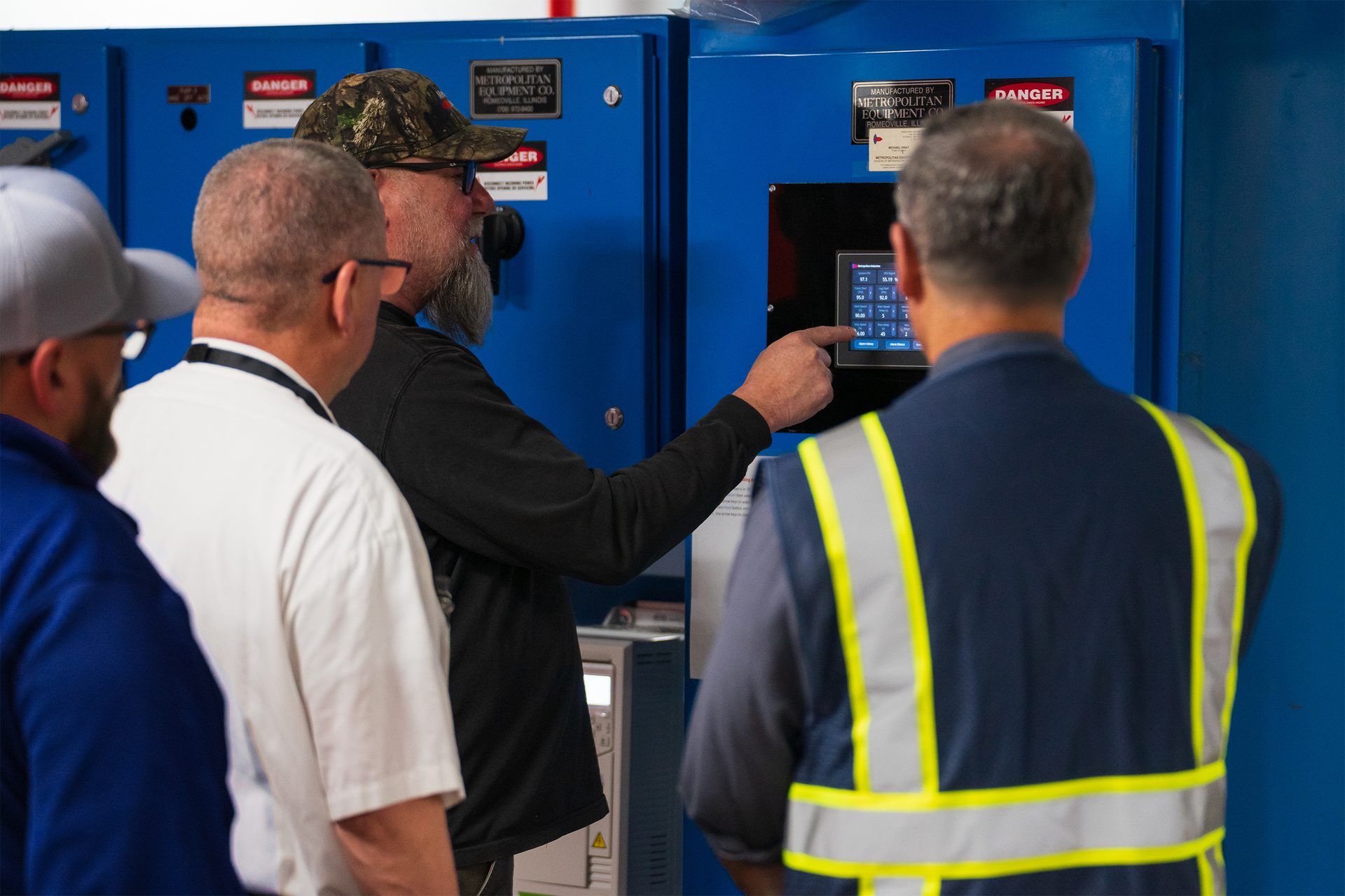 Man points at a digital control panel on blue industrial equipment while others observe.