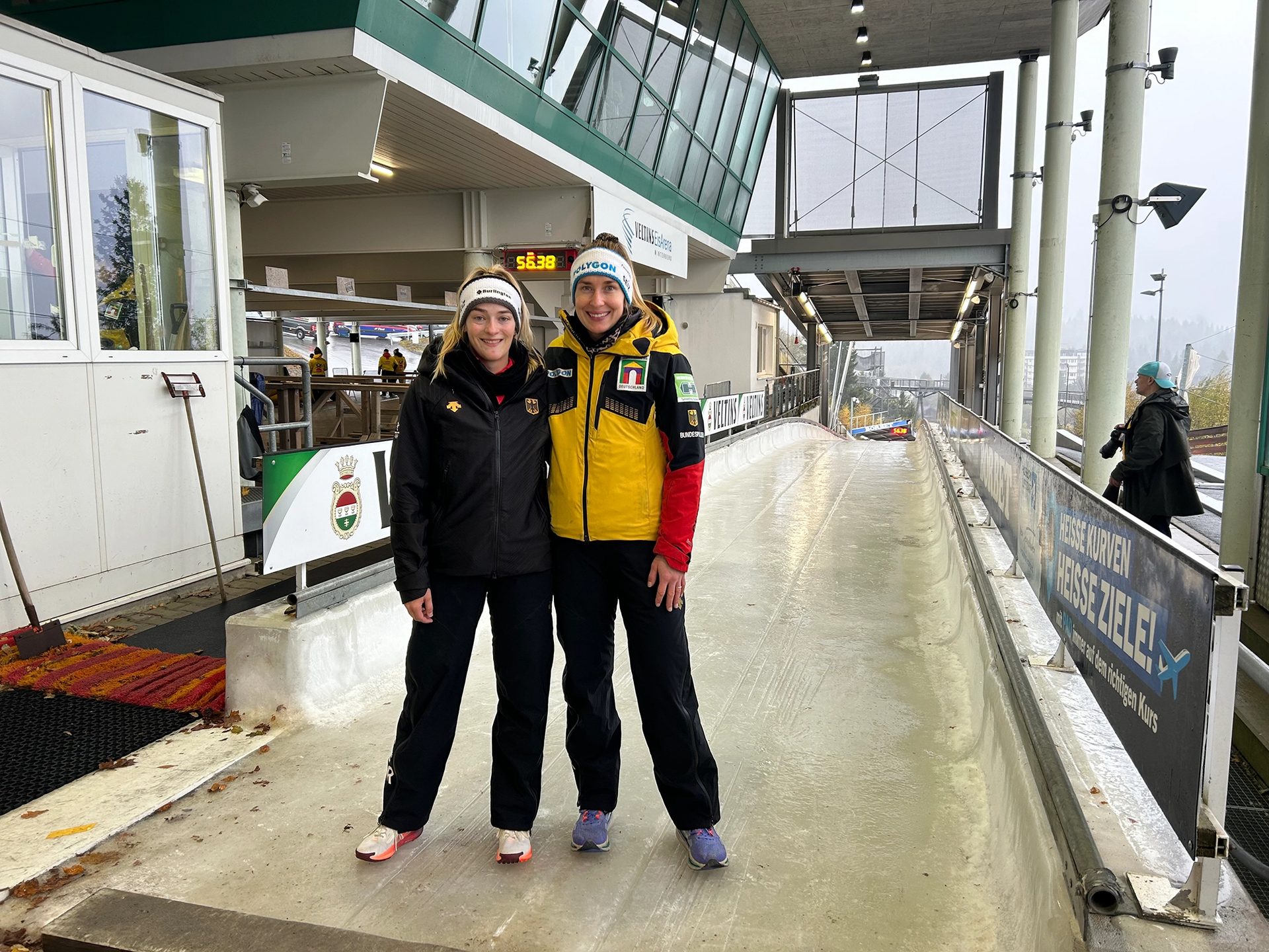 Two smiling female athletes in winter gear on an ice track at VELTINS EisArena, with a timer reading 56.38.