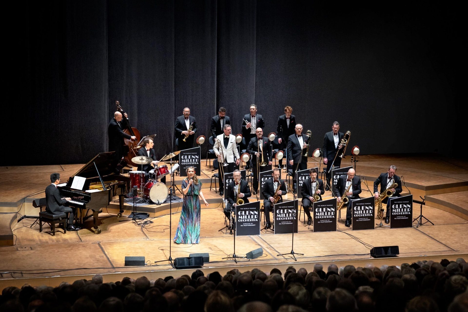 The Glenn Miller Orchestra performs live on stage with a singer, piano, brass, and woodwinds.