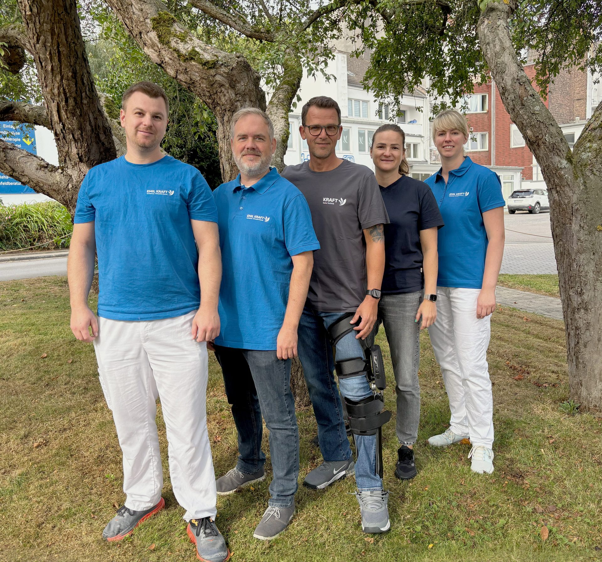 Five healthcare professionals, one with a leg brace, stand outdoors near a tree.