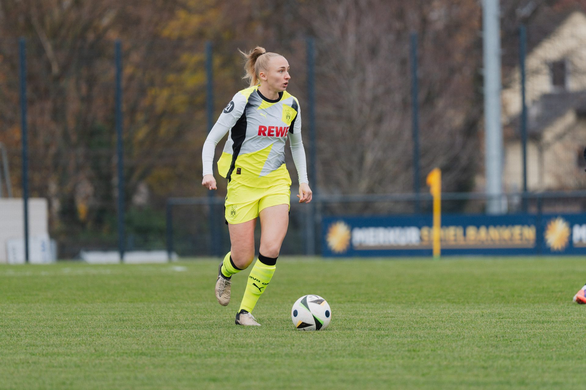 Female soccer player in a yellow and gray uniform dribbling a ball on a green field.