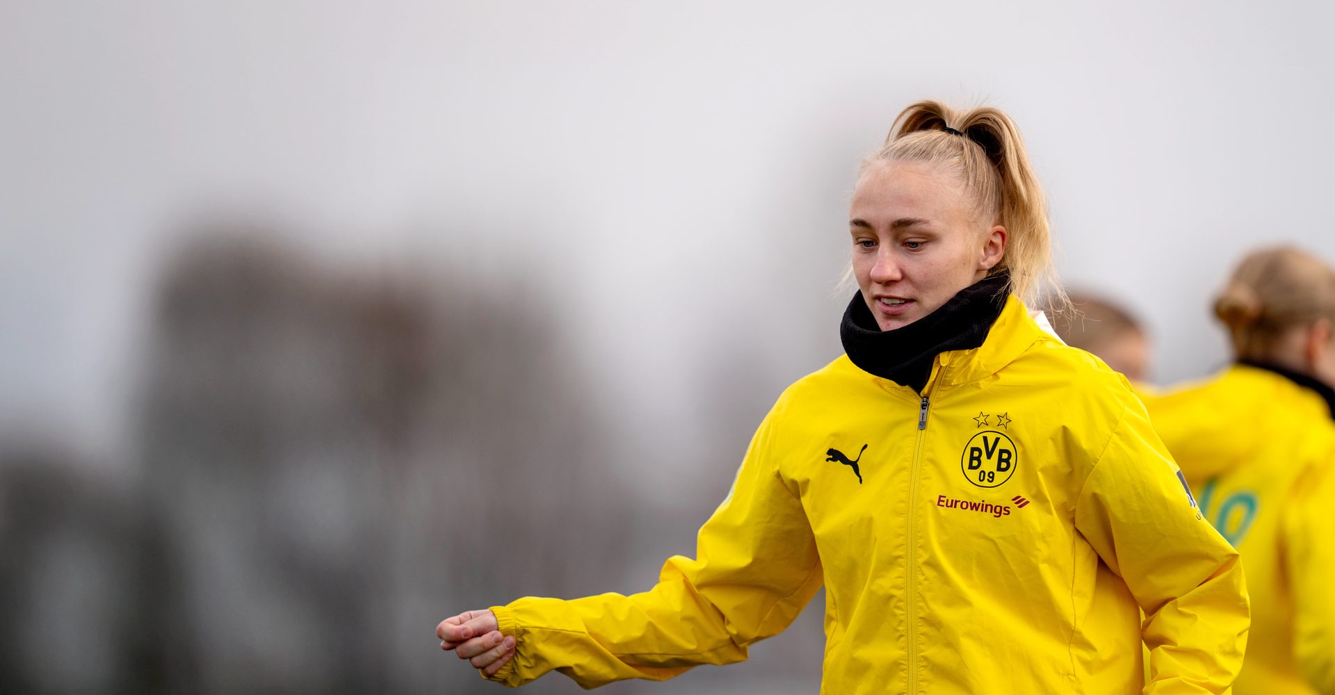 A woman in a yellow Borussia Dortmund jacket with a ponytail looks right, outdoors.