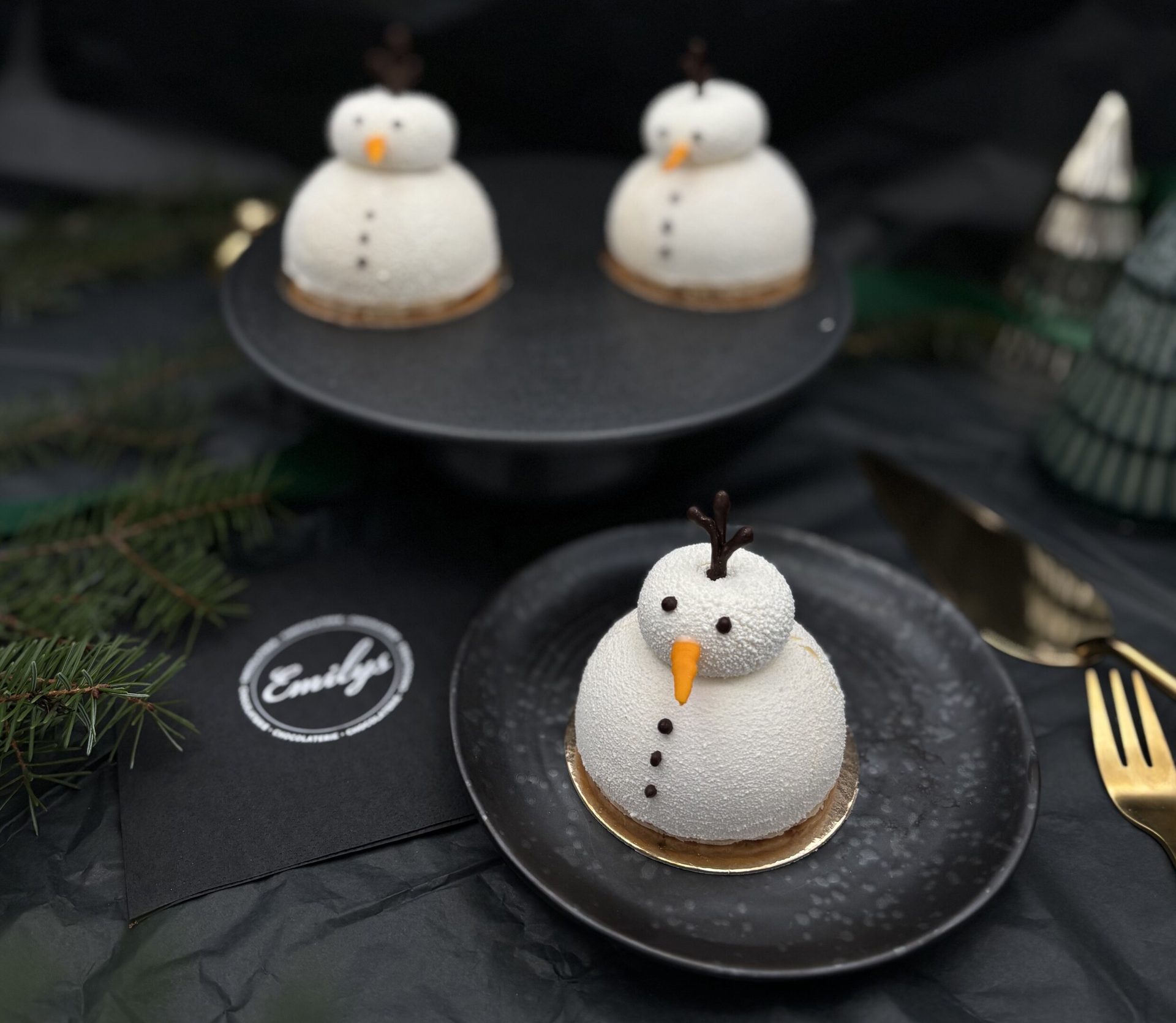 Three white snowman desserts on black plates, with pine branches and winter decorations.