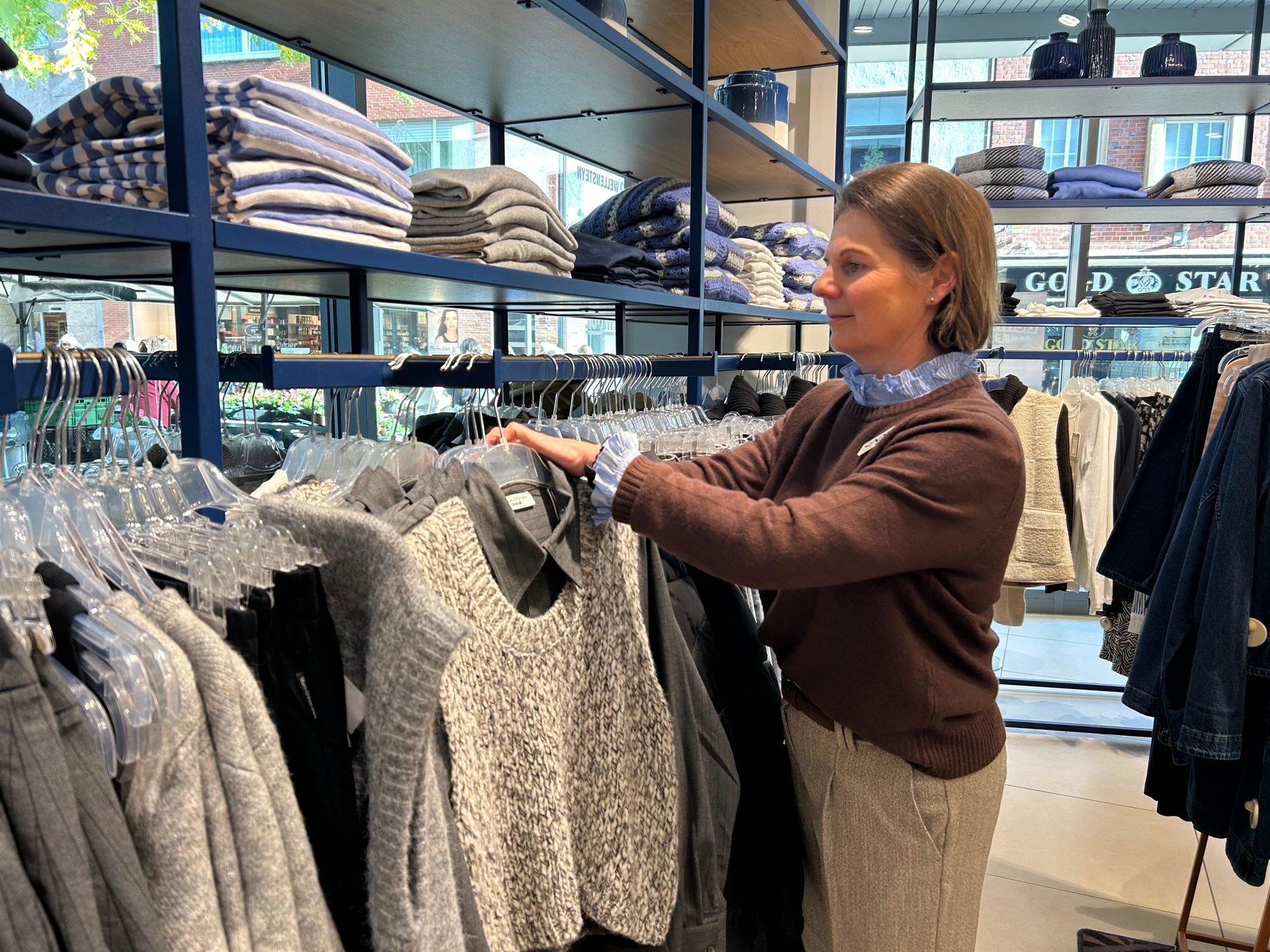 A woman in a brown sweater browses clothes on a rack in a retail store.