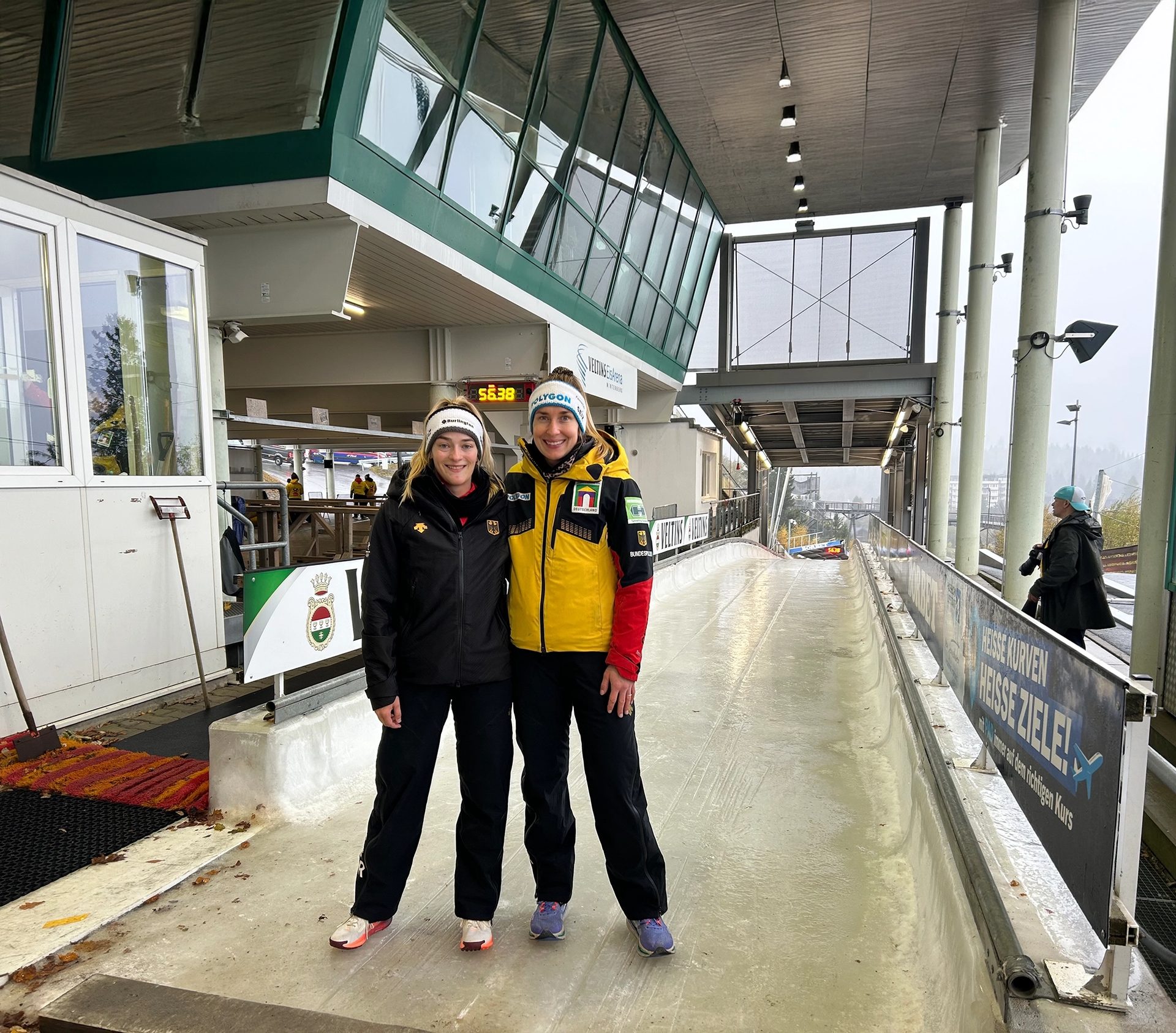 Two female athletes in winter gear on a bobsled track with a start building in the background.