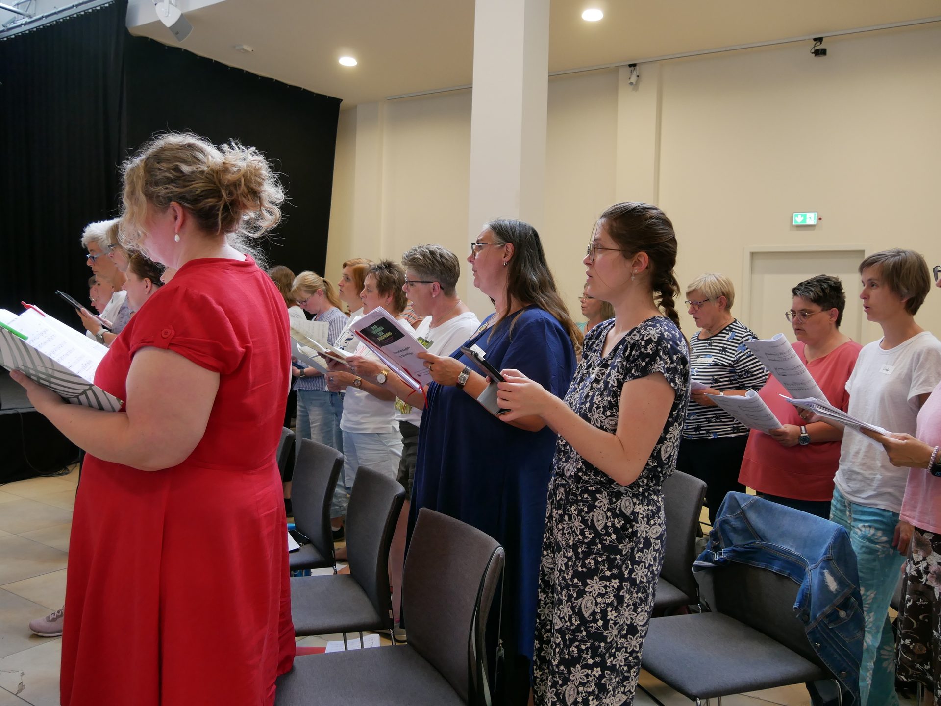 Women sing in a choir, holding sheet music, practicing in a well-lit room.