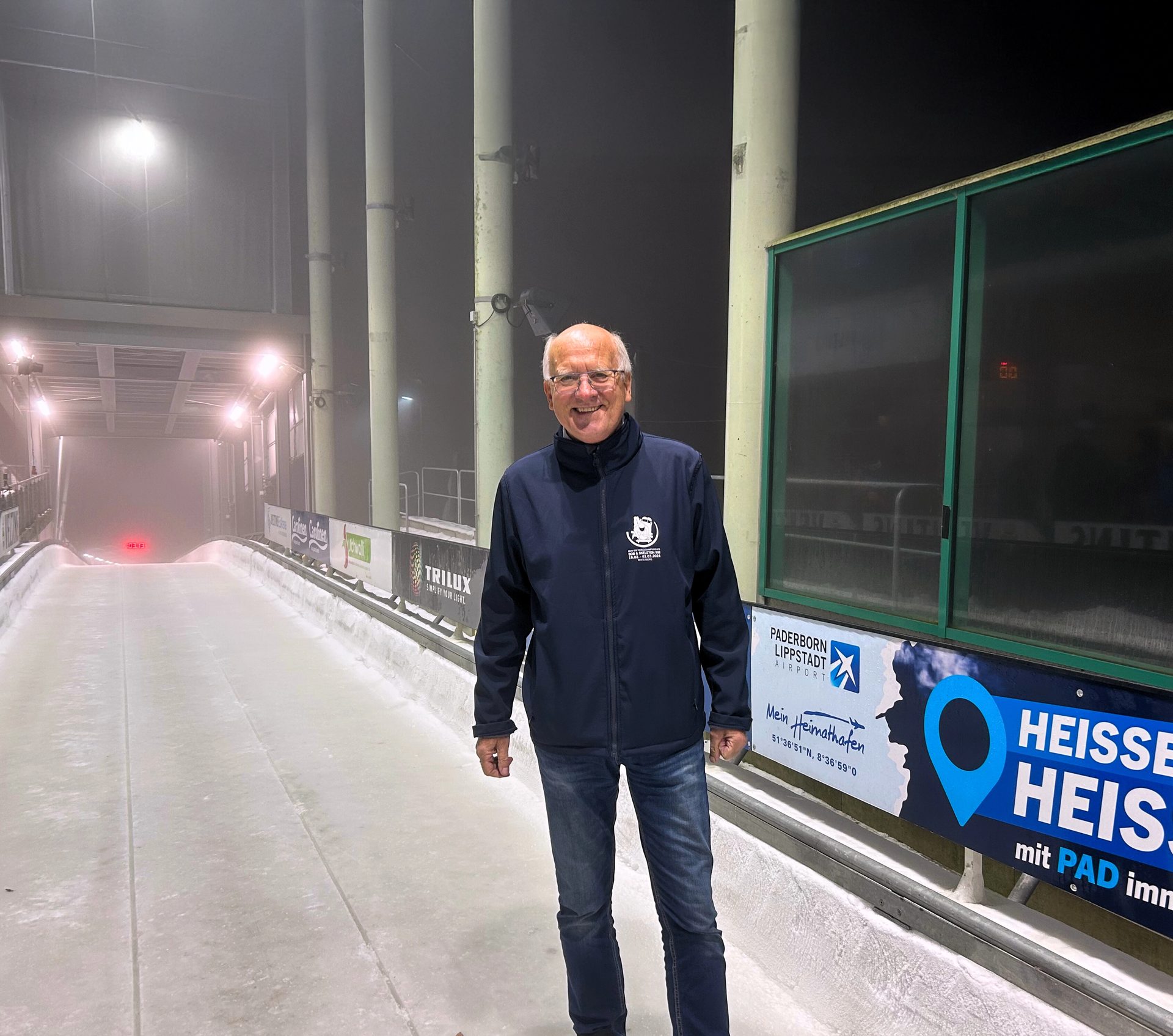 Smiling man on an illuminated ice track at night, with sponsor banners in the background.