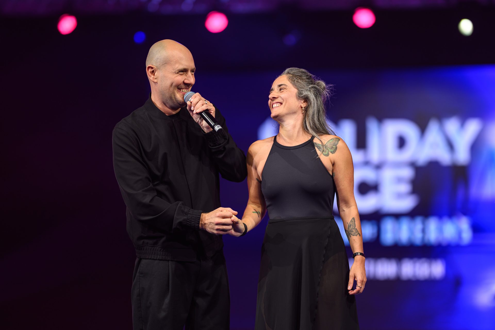 Smiling bald man with mic and woman with gray hair holding hands on stage.