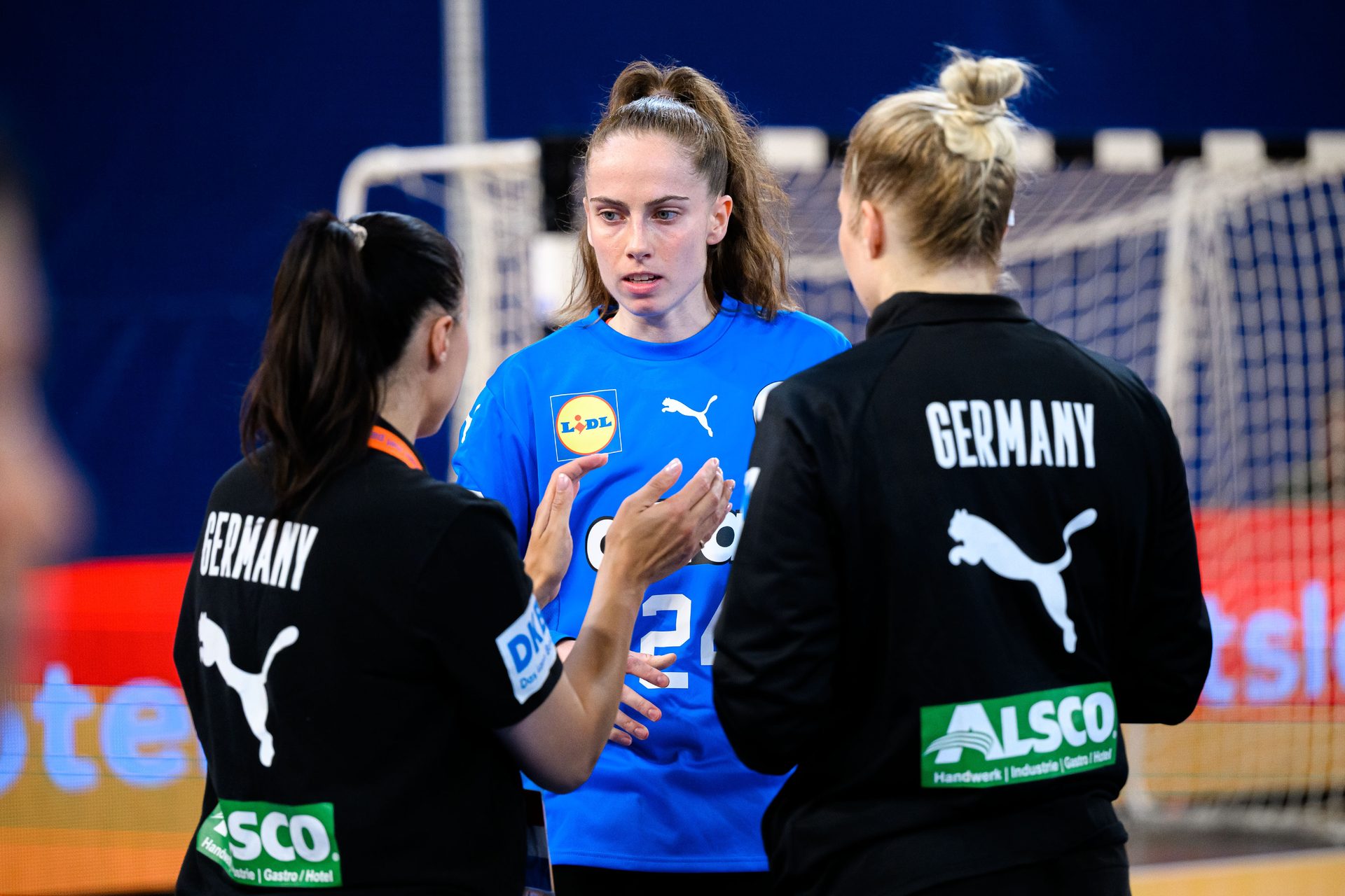 Three female handball players: two with "GERMANY" on their backs, one in blue talks and gestures.
