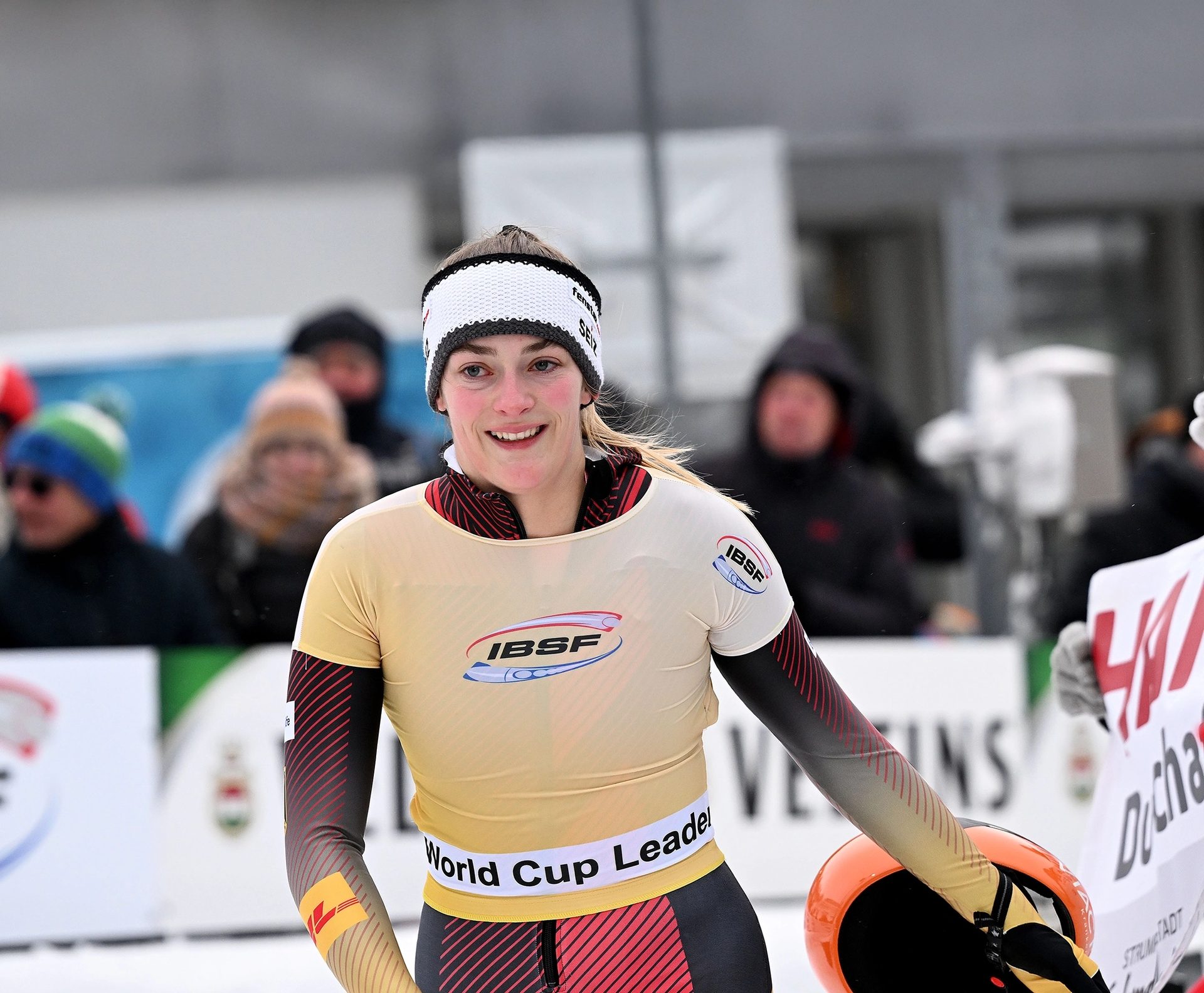 Smiling female athlete in IBSF World Cup Leader suit, headband, holding a helmet.
