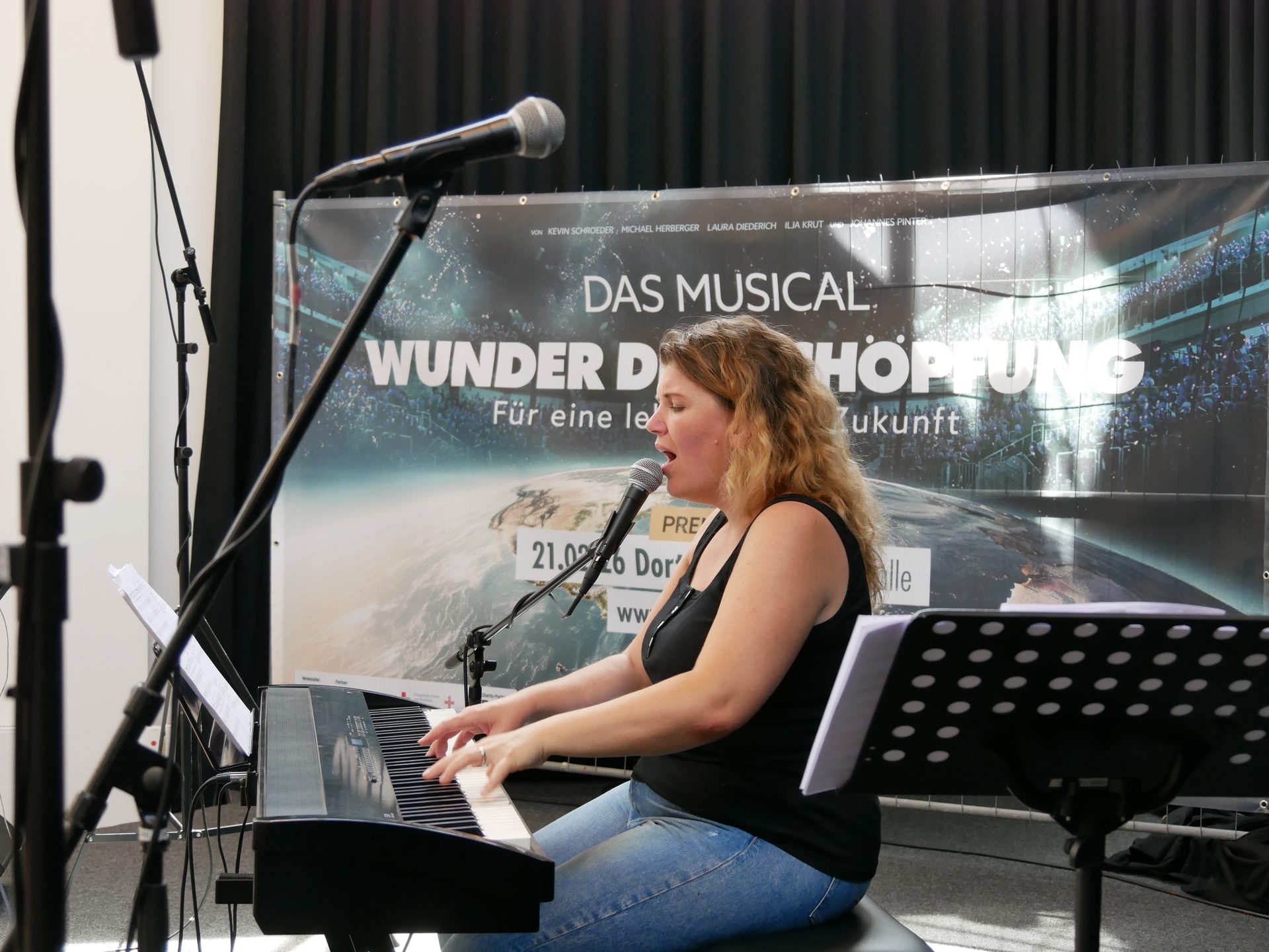 Blonde woman singing into a microphone and playing a digital piano in front of a "Das Musical" banner.