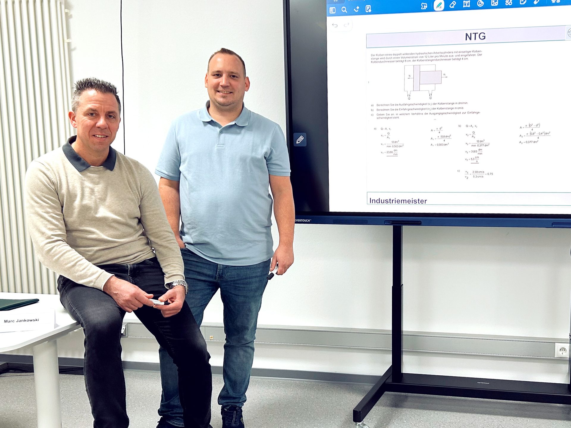 Two men, one sitting and one standing, in front of a large interactive screen displaying a math problem.
