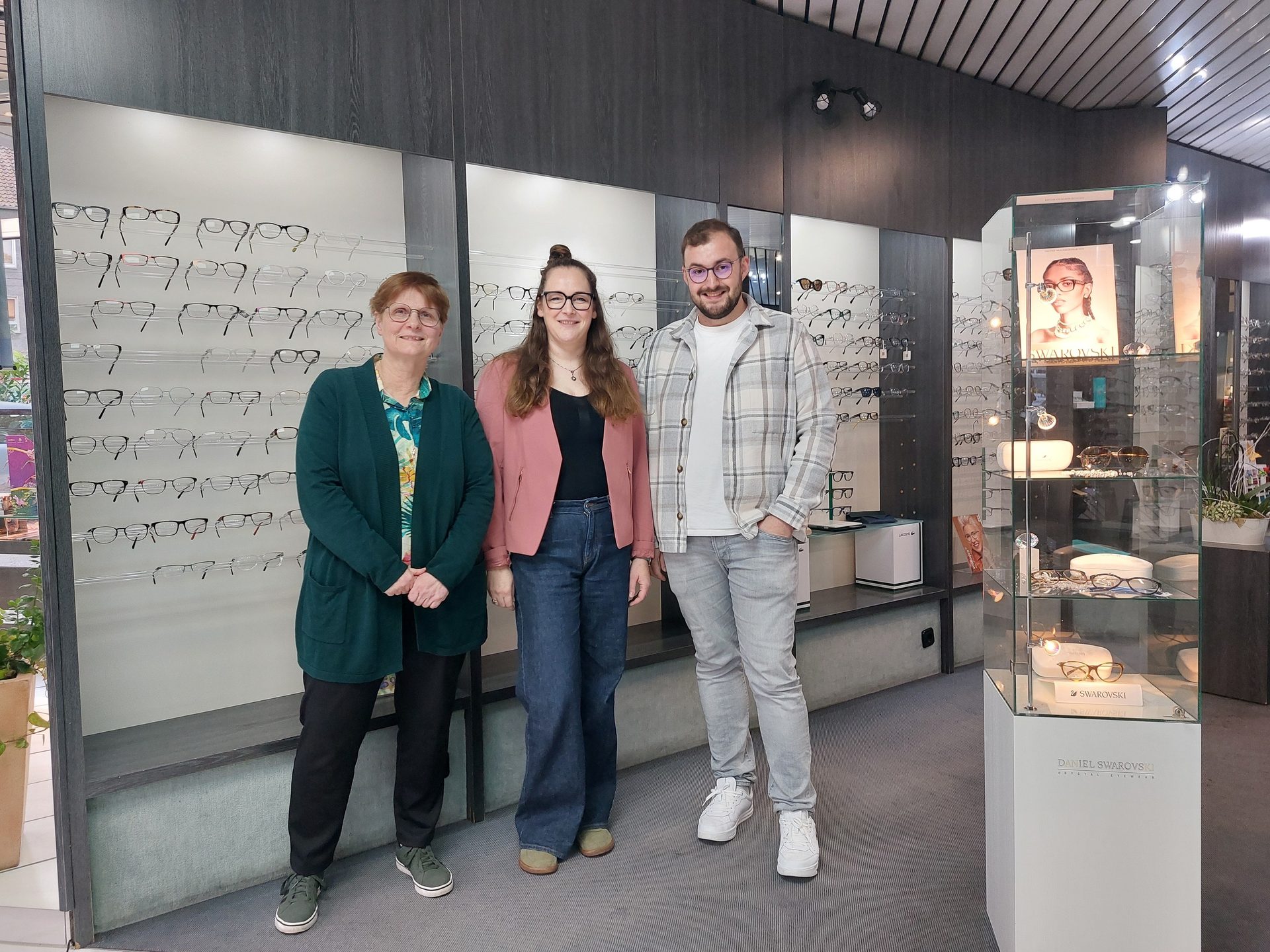 Three smiling people in an optician's store surrounded by glasses displays.
