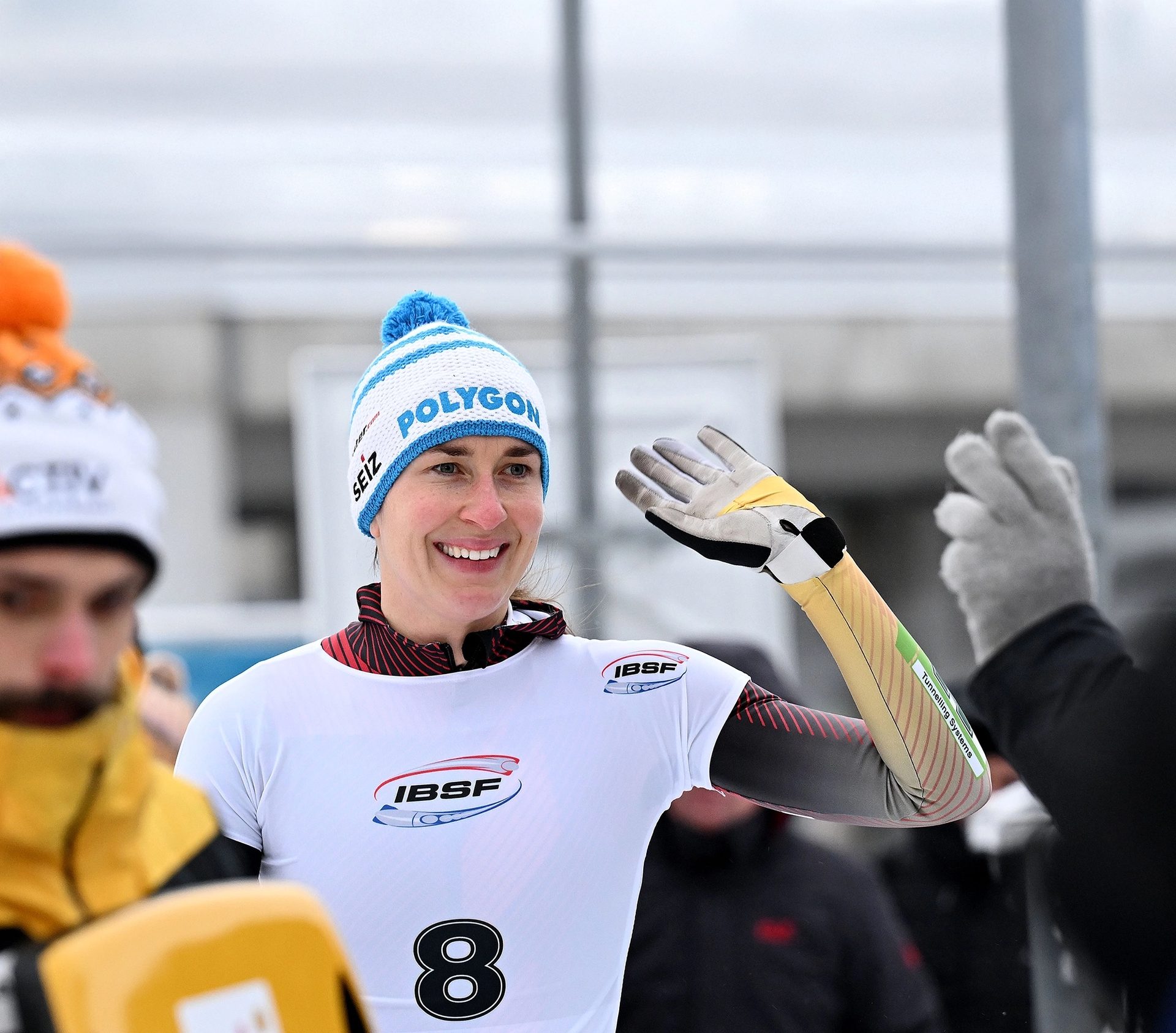 A smiling woman in a 'Polygon' beanie and winter sports uniform waves her hand.