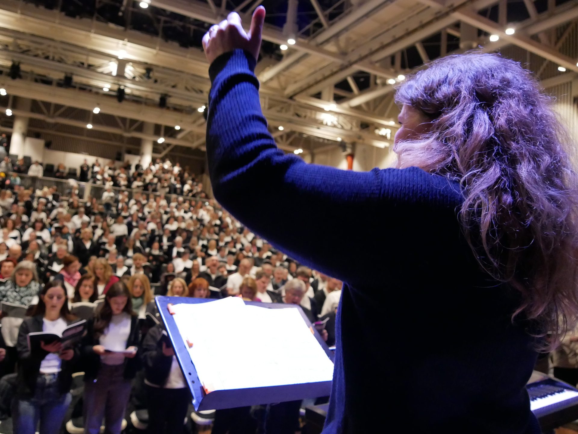 Female conductor, seen from behind, arm raised, leads a large choir with a music stand.