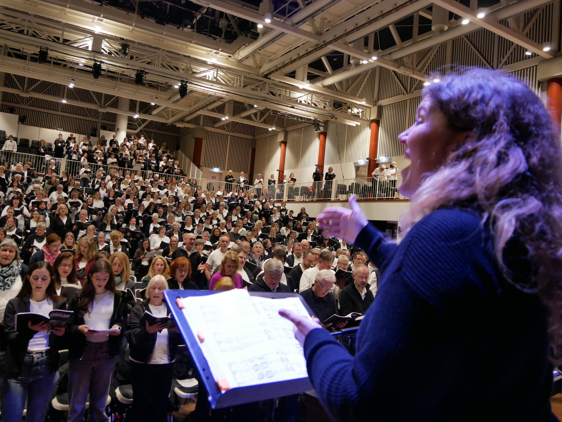 A choir conductor directs a large choir in a concert hall, with many singers holding sheet music.