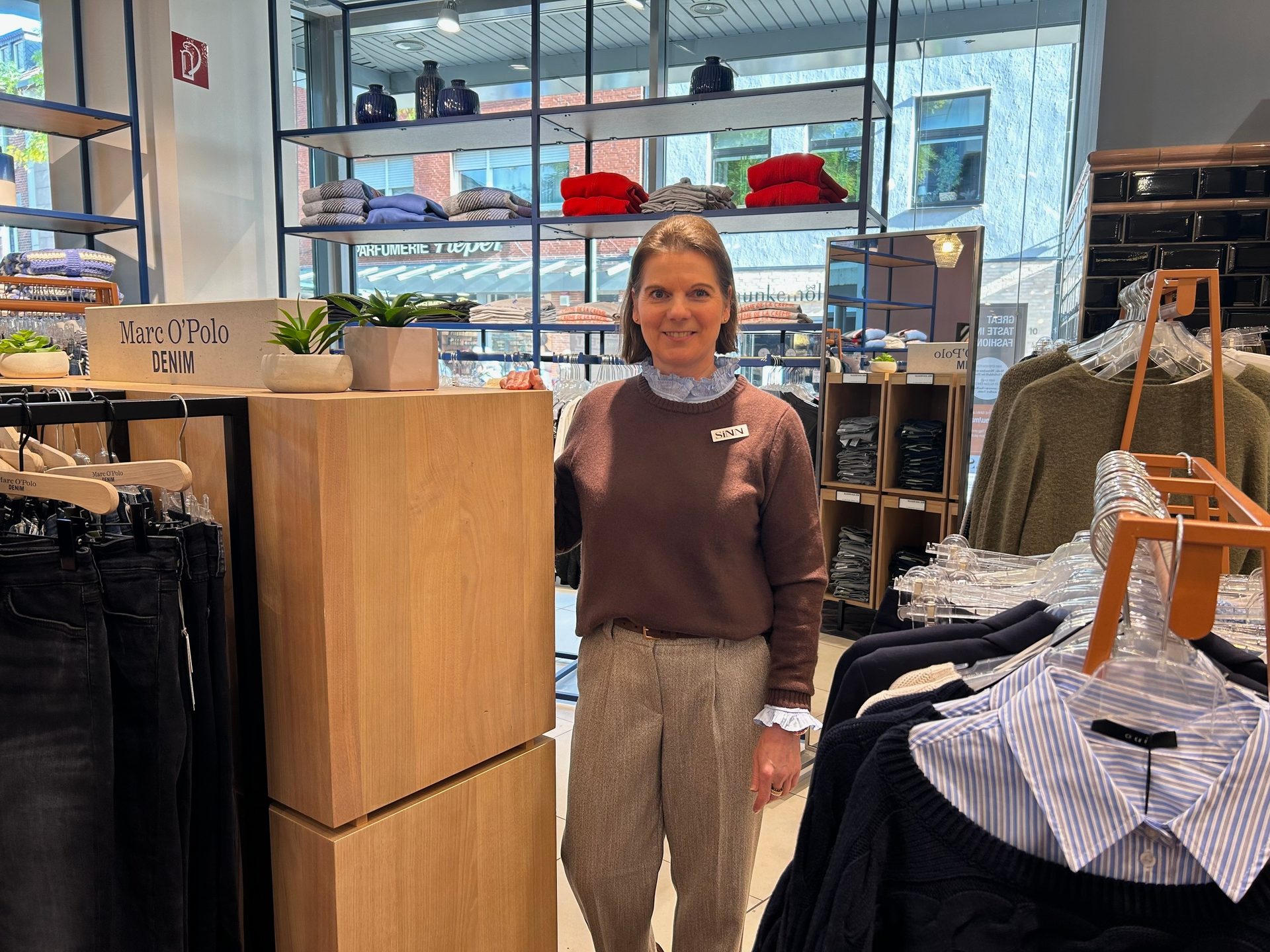 Smiling woman in a brown sweater and plaid pants in a Marc O'Polo clothing store.