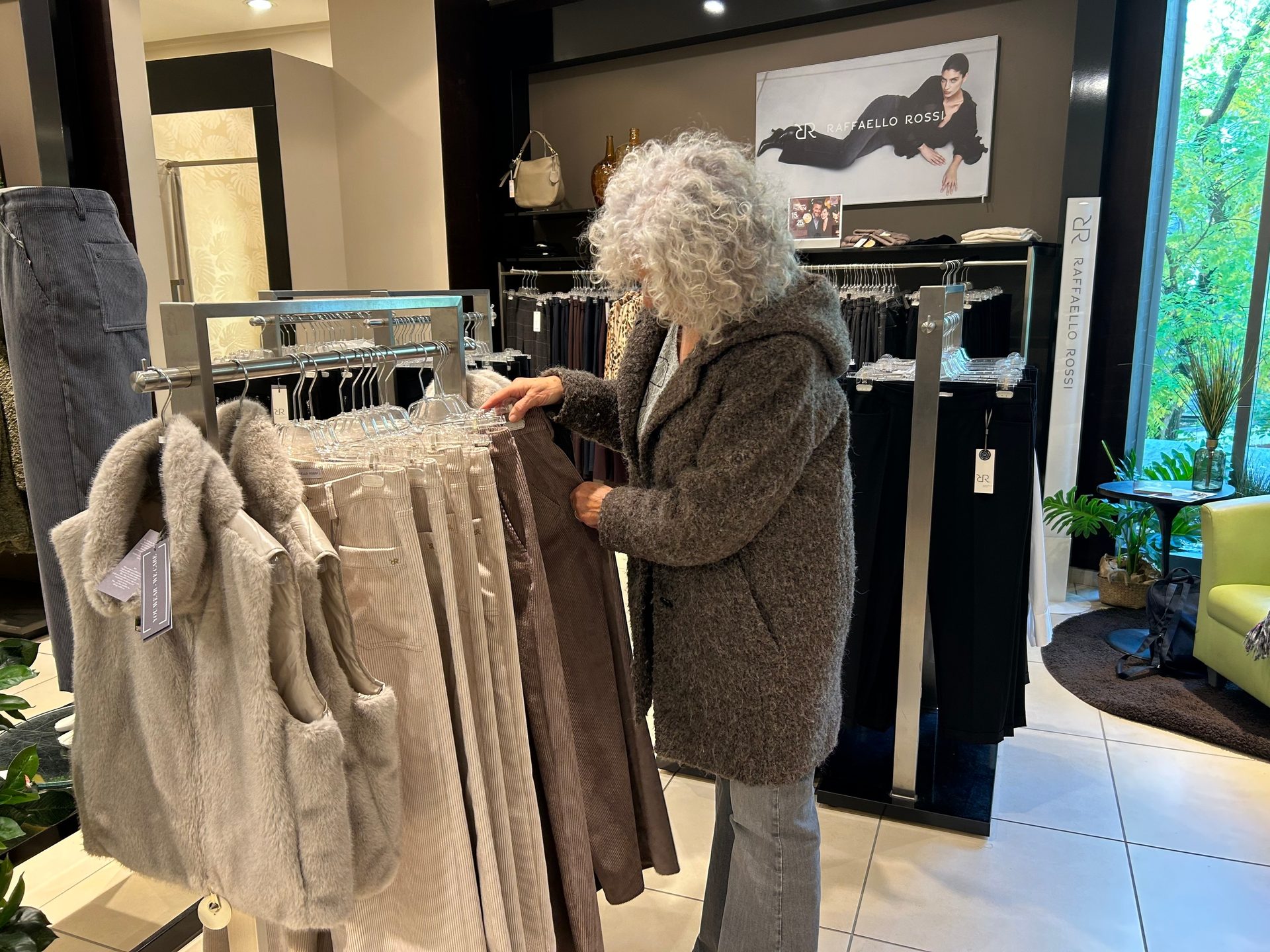An older woman with curly white hair browsing clothes on a rack in a store.